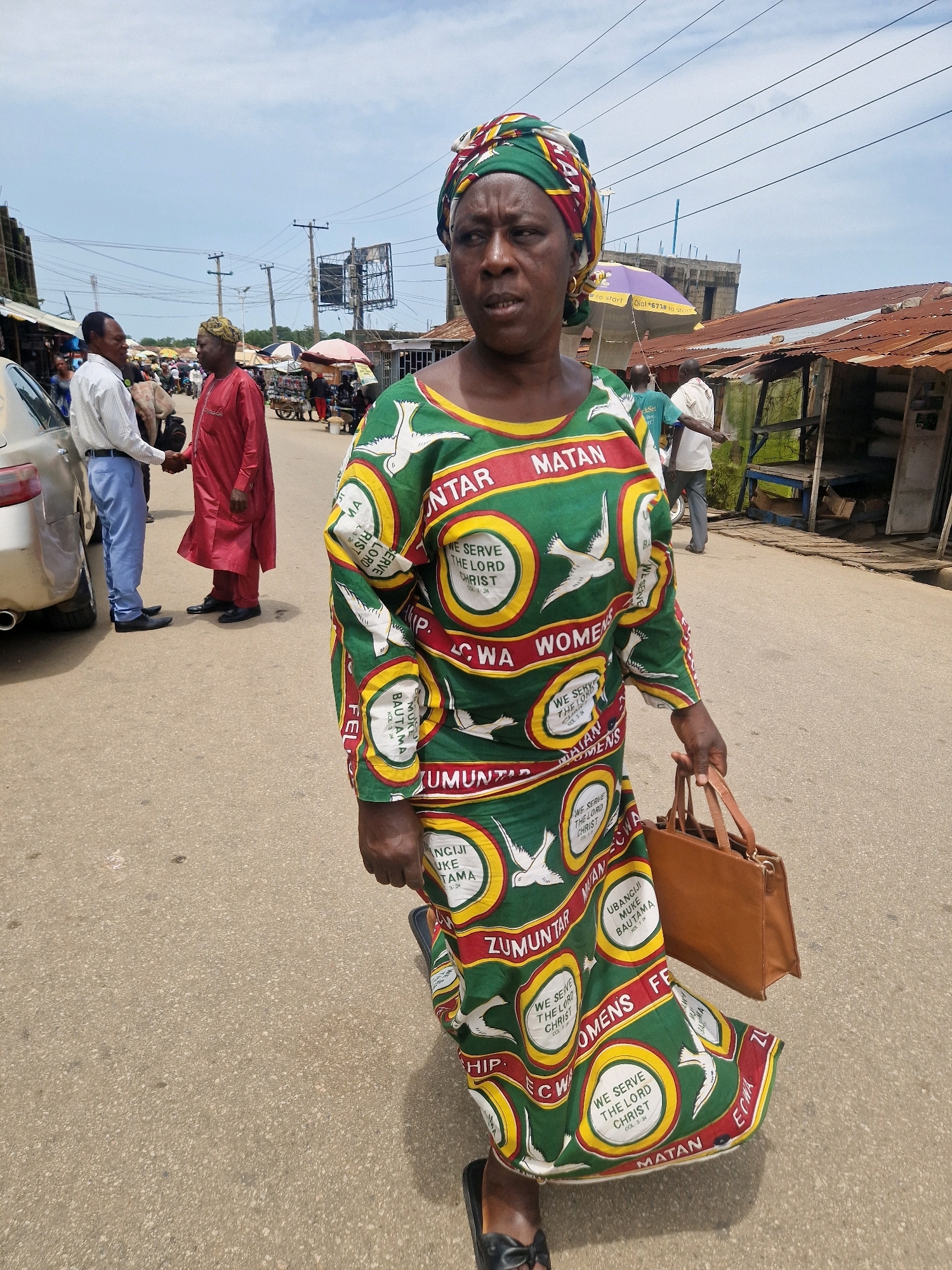 In an outdoor market scene, a woman in a colorful headscarf and a green dress with white bird motifs and text walks towards the camera, carrying a brown handbag. She is in the foreground, with other market-goers and stalls visible in the background. Two men are shaking hands on the left side of the image, while several market stalls with corrugated tin roofs and umbrellas are set up on the right. The ground is paved with asphalt. The sky is partly cloudy.