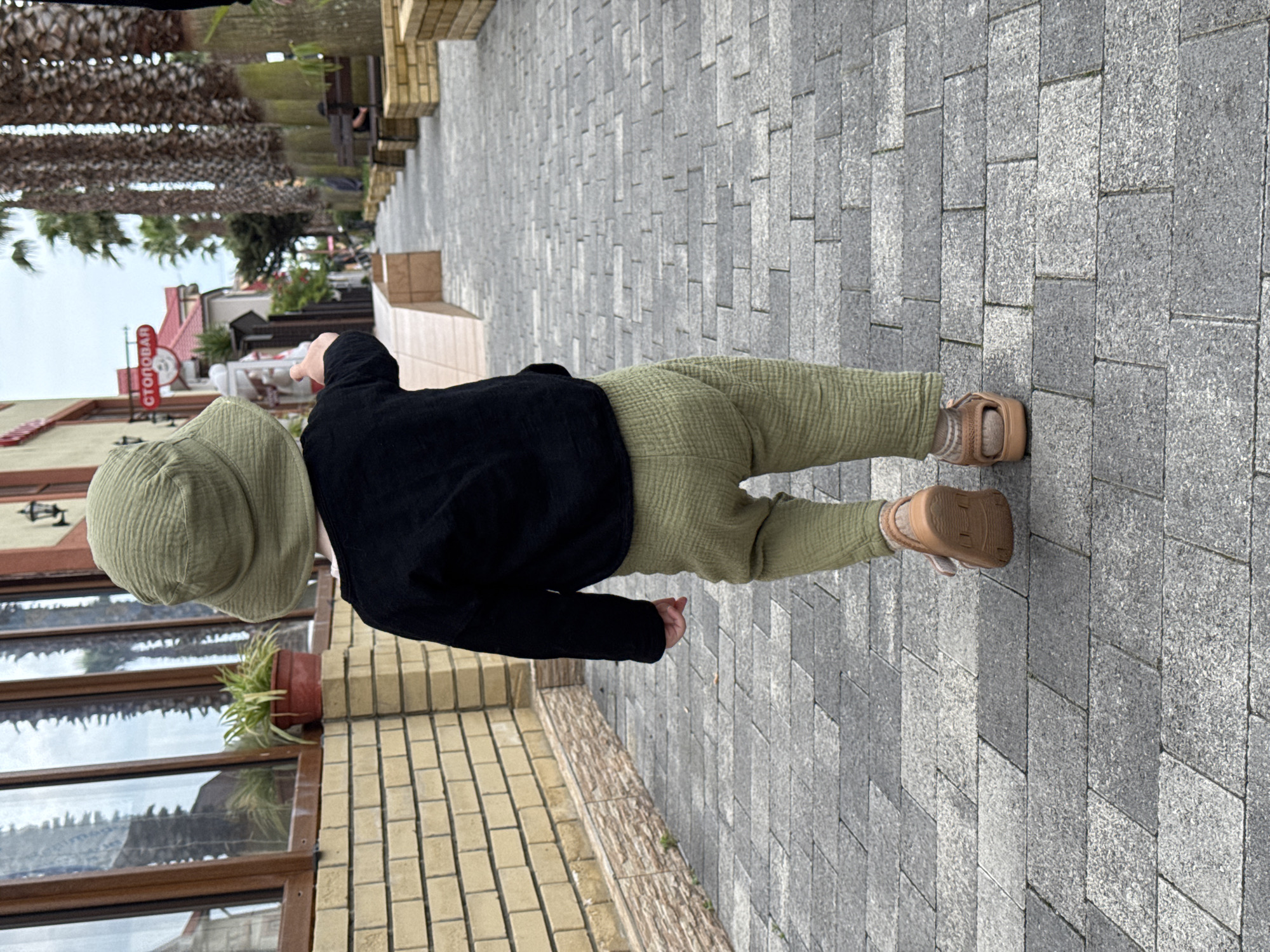 A child wearing a green hat, a black shirt, green pants, and tan sandals is walking on a herringbone-patterned paved sidewalk in the city of Sirius, Russia. The child is walking near a building with windows and a potted plant. A sign with Cyrillic writing is visible in the background. Palm trees are also visible in the background.
