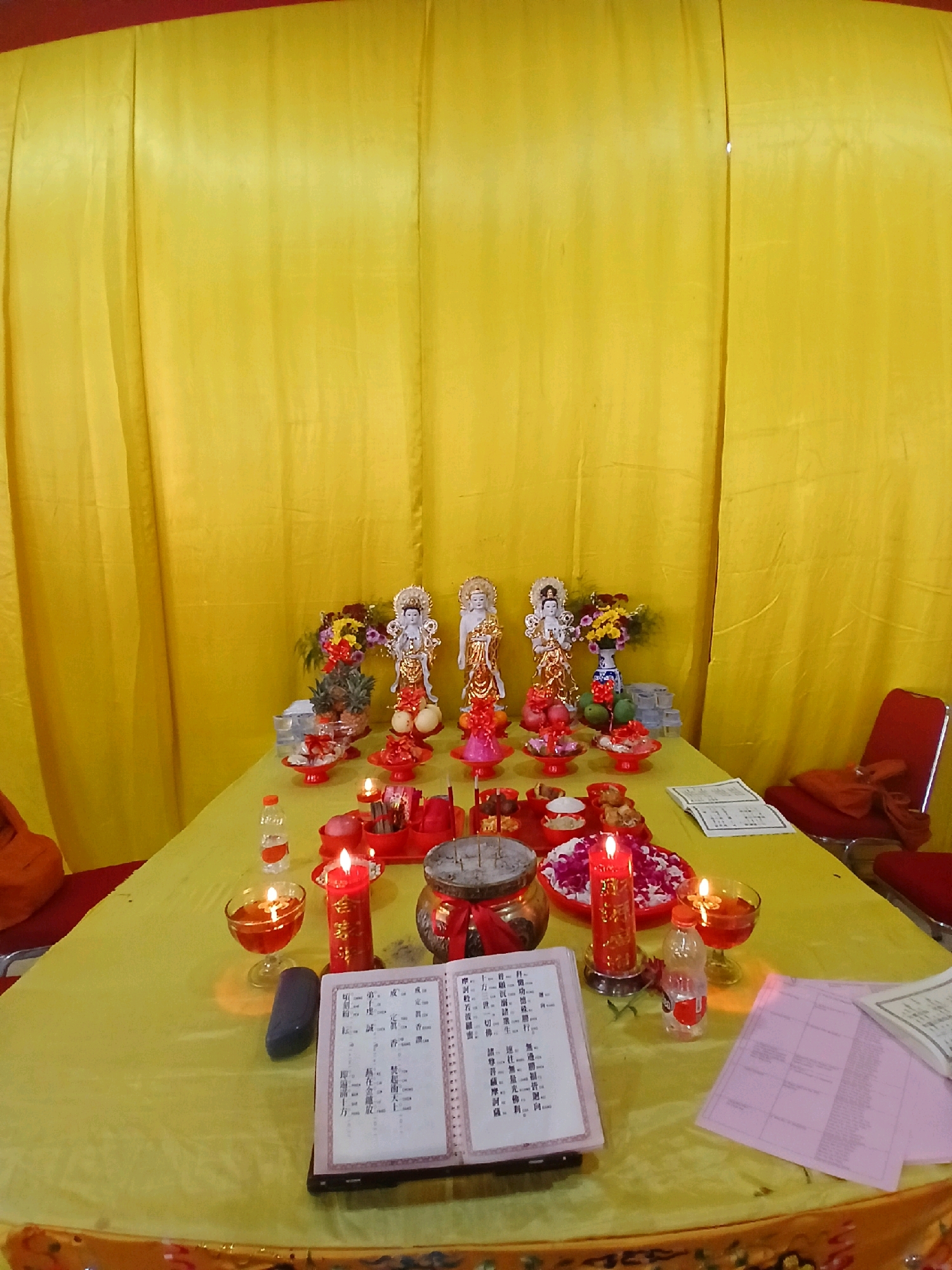 In an interior setting with a vibrant yellow curtain backdrop, a table is covered with a light green tablecloth, upon which a variety of items are arranged for what appears to be a religious or ceremonial offering. Dominating the scene are three statues of figures, possibly deities or revered individuals, adorned in ornate clothing and elaborate headdresses. These statues are positioned behind a collection of offerings, including fruits such as oranges and what appear to be pineapples and mangoes, arranged in red bowls.
Several red candles, some lit and casting a warm glow, are placed around the table. One prominent red candle stands tall on a silver holder, while another is smaller and shorter. There are also small glasses filled with a reddish liquid, possibly incense or oil, which are also lit, creating a gentle luminescence. In the foreground, an open book with Chinese characters is displayed on a black stand, suggesting a ritualistic text or prayer book. Next to it, a dark, elongated object, possibly a prayer bead or a decorative stone, rests on the tablecloth.
Other objects on the table include a lidded pot with a red ribbon, a small red bowl filled with white and pink flower petals, and a clear plastic bottle of water. In the background, a red chair with an orange cloth draped over it is visible, hinting at a place for a participant or attendee. The overall atmosphere suggests a solemn and respectful occasion, with the offerings and candles creating a serene ambiance. The specific location is identified as Unknown, Unknown.