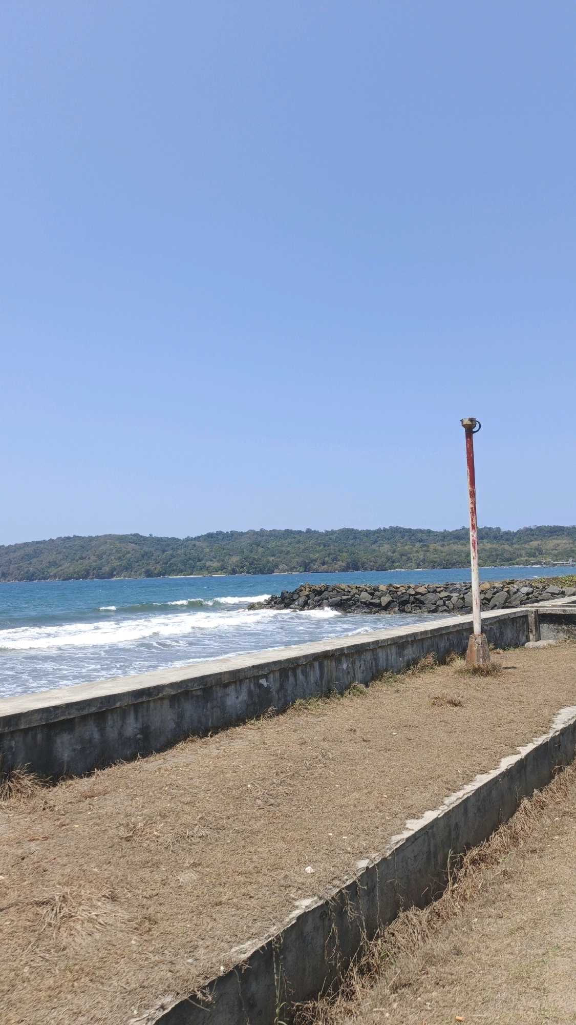 The image shows a view of a beach and the ocean, with a clear blue sky overhead. In the foreground, there is a concrete wall bordering a dry, grassy area. A red and white striped pole with a metal fixture at the top stands in the grassy area to the right. Beyond the wall and grass, the ocean meets the shore with gentle waves. A rocky breakwater extends into the water, and a tree-covered hillside forms the background. The scene is static and appears to be a sunny day in an outdoor coastal location. No people or animals are visible. The location is identified as Desa Pagandaran, Indonesia.