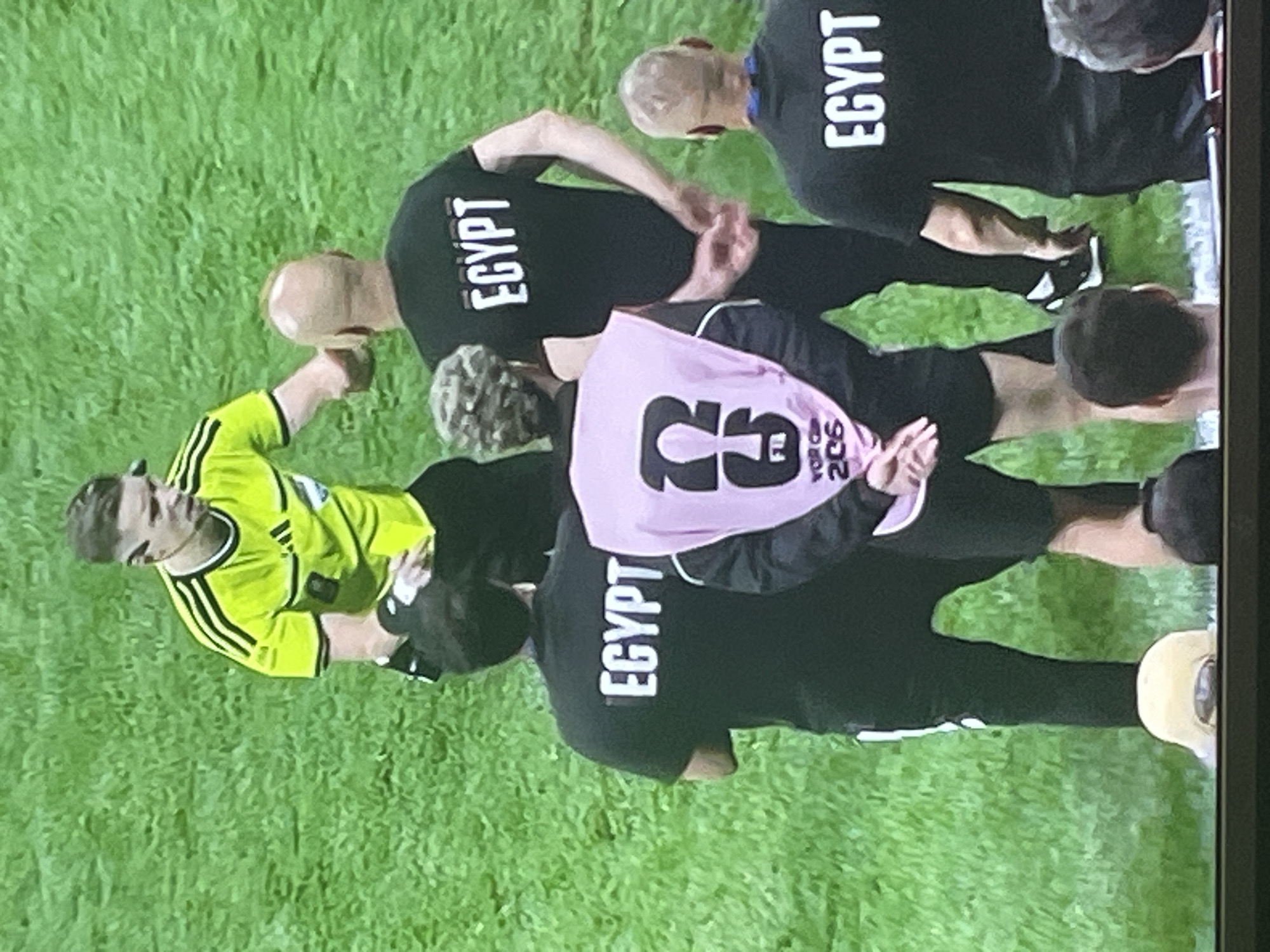 The image shows a group of people on a green, grassy field, likely a sports field. A person in a bright yellow referee's shirt with black stripes on the sleeves is interacting with several other individuals. Some of these individuals are wearing black shirts with "EGYPT" written on them in white. One person in the foreground is wearing a pink vest over their clothing, which has a large number "2" on it along with some text. The scene appears to be from a sporting event. The location is not Zhukovo, Russia, but rather an unspecified outdoor sports venue.