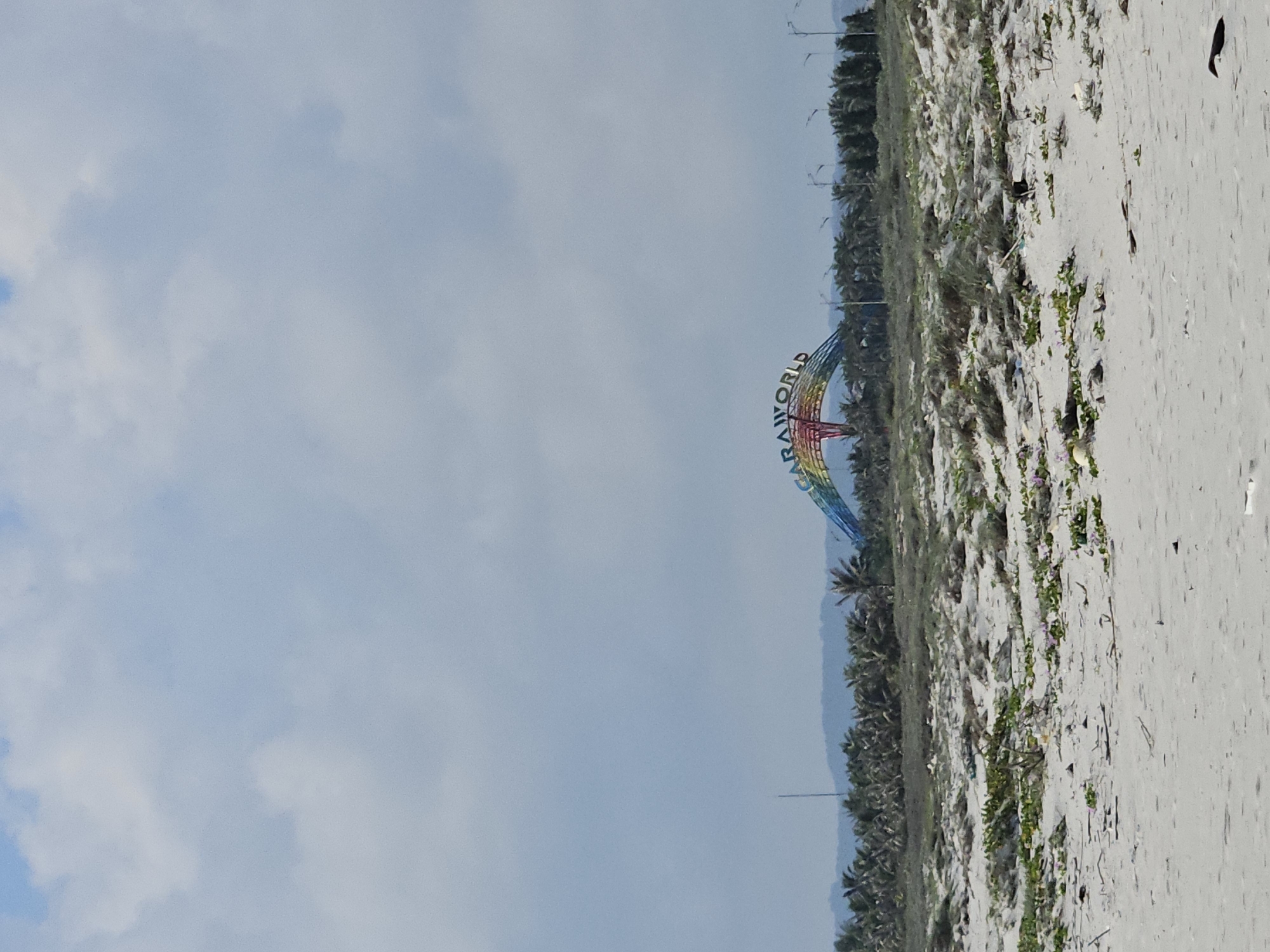 The image displays a coastal landscape under a partly cloudy sky. A wide expanse of light-colored sand, possibly a beach or dune area, occupies the foreground and mid-ground. Scattered across the sand are patches of green vegetation and some natural debris or litter.
In the mid-distance, a prominent, colorful archway structure stands, featuring a rainbow design. Clearly visible on this archway is the text "SEAWORLD," indicating an amusement park or marine attraction. Behind the archway and extending along the horizon to the left, a line of numerous palm trees suggests a tropical environment. Beyond the trees, a faint, hazy landmass or distant shoreline can be seen.
The sky is a pale blue, adorned with scattered white and light grey clouds, suggesting an overcast or partly sunny daytime. No people or specific activities are visible in the scene. The overall impression is of a quiet, possibly undeveloped beach area adjacent to a themed attraction. Given the "SEAWORLD" sign and tropical foliage, the setting aligns with a coastal resort or park, consistent with the provided location context of Bac Cam Ranh Ward, Vietnam.