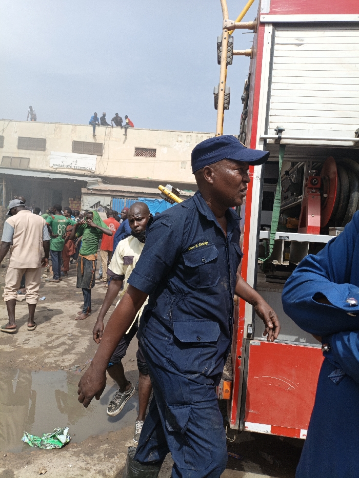 This image captures an outdoor scene in Fagge C, Nigeria, depicting a crowd of individuals and an emergency vehicle. In the foreground, a male individual, identified by a name tag as "Usman B. Zangina," wears a dark blue uniform consisting of a collared shirt with two breast pockets and matching trousers, along with a dark blue baseball cap. He stands facing right, his right arm partially visible. To his immediate right, the red and white side of a large fire truck is visible, featuring a white roll-up compartment door and a black hose reel. A yellow metallic structure, possibly part of an extendable ladder or crane, extends upwards from the truck.
In the mid-ground, behind the uniformed individual, a diverse crowd of men is gathered on an unpaved, muddy ground surface that includes puddles. Several men wear light-colored shirts, while others are in green shirts, one displaying the number "83." One man in a yellow shirt is partially obscured. Further in the background, a light-colored, flat-roofed building bears a sign reading "MAZAF UTC TECHNICAL," with additional letters "COMP" partially visible on the building's facade. Five individuals are seated atop this building, observing the scene below. The sky above is clear and bright. On the ground in the immediate foreground, a crumpled green object lies near a puddle.