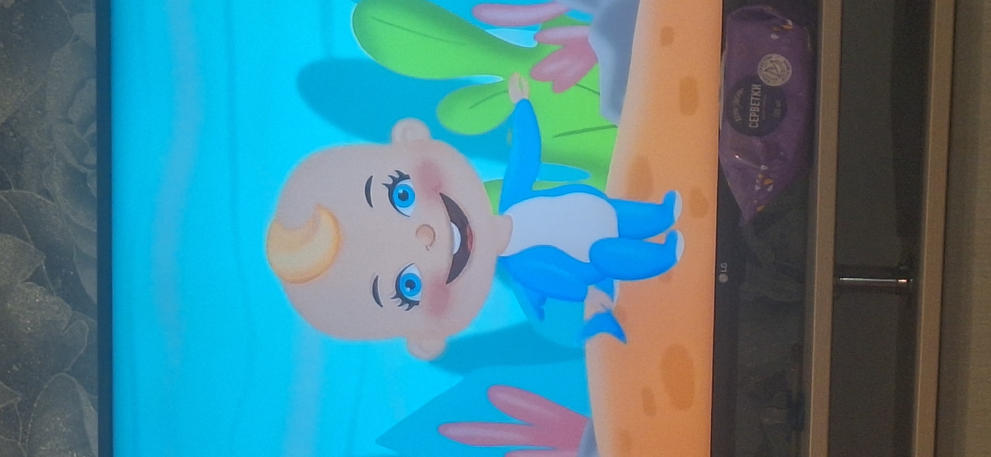 The image shows a television screen displaying a cartoon character that appears to be a baby wearing a shark costume. The character has a round head with light blonde hair, large blue eyes, and rosy cheeks. It is smiling broadly and appears to be standing on an orange surface. In the background, there are stylized underwater plants, suggesting an aquatic environment. The TV screen itself is mounted on a stand, and at the bottom of the screen, there is an LG logo. On the stand, below the TV, is a purple package of wet wipes with Russian text that translates to "SERBETKI" (wipes). The scene is viewed from a slight angle, and the lighting suggests it is indoors. There is no information in the image to indicate its location in the city of Bila Tserkva, Ukraine.