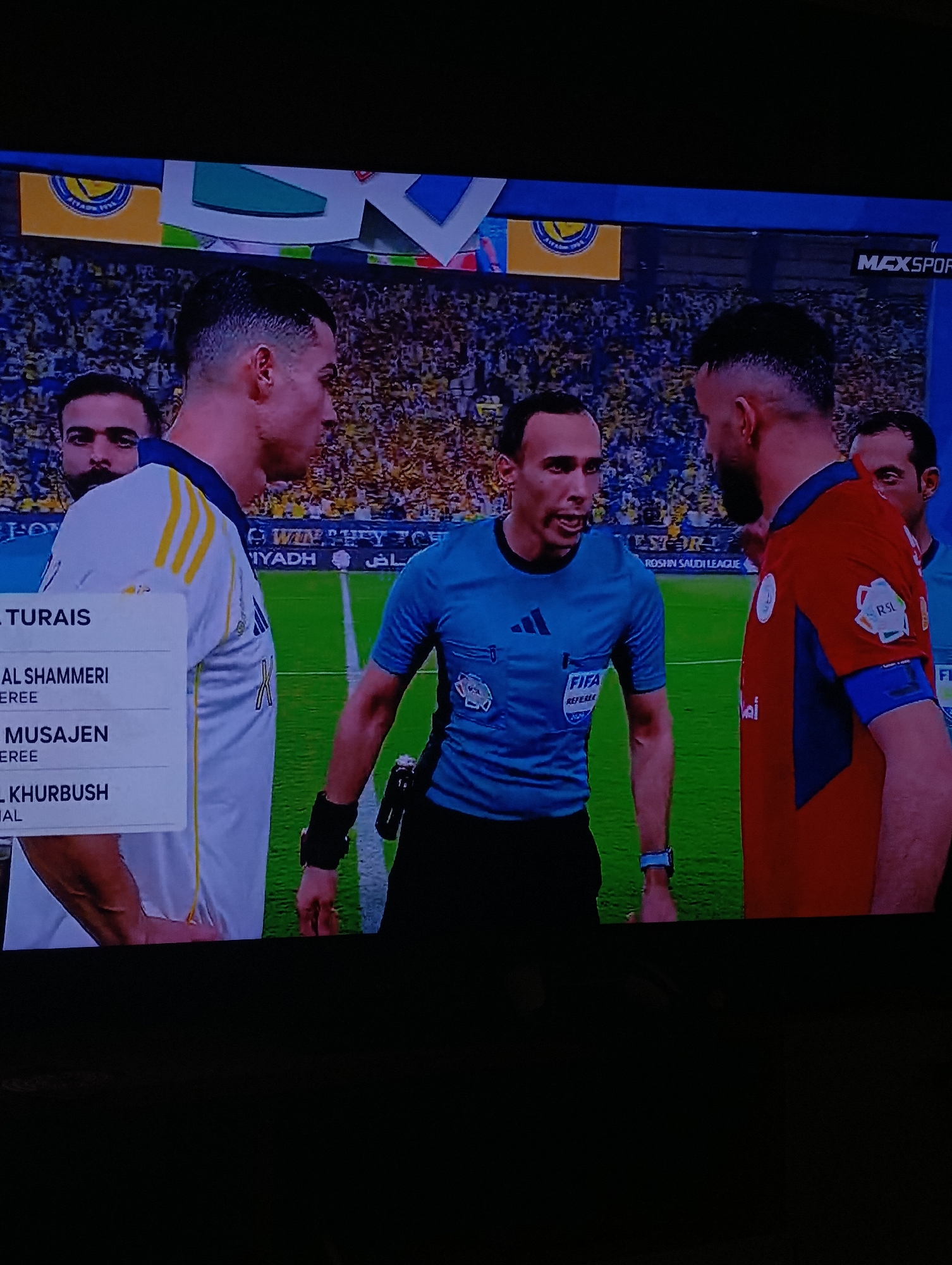 The image shows a scene from a soccer game, likely a professional match. Two soccer players, one in a white and yellow jersey and the other in an orange and blue jersey, are in a face-to-face confrontation. A referee wearing a blue FIFA referee shirt stands between them, seemingly mediating the situation. Other players are visible in the background. The game appears to be taking place in a stadium with a crowd of spectators. Text on the screen indicates the “ROSHIN SAUDI LEAGUE” and the names of the match officials. MAXSPORT logo also can be seen. The game is not in the city of Plovdiv, Bulgaria.