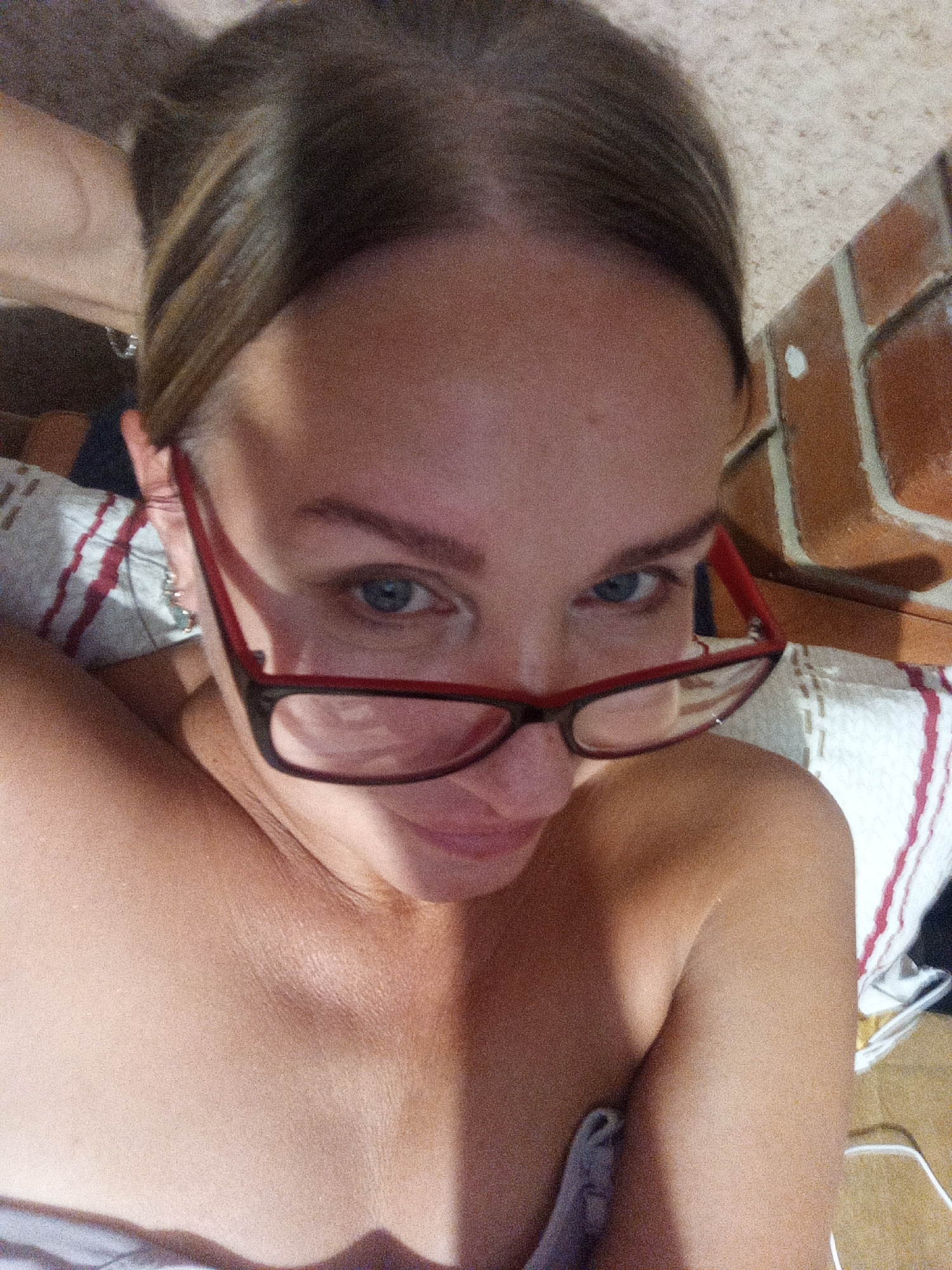 A medium shot, eye-level, indoor selfie shows a woman with light skin and brown hair. She is wearing red-rimmed glasses and looking directly at the camera. The woman is lying down, propped up on a striped pillow that has red and white stripes. To the right of the woman, there is a brick wall with visible grout lines. The lighting in the image suggests it was taken with artificial light. The overall scene appears to be in a domestic setting.