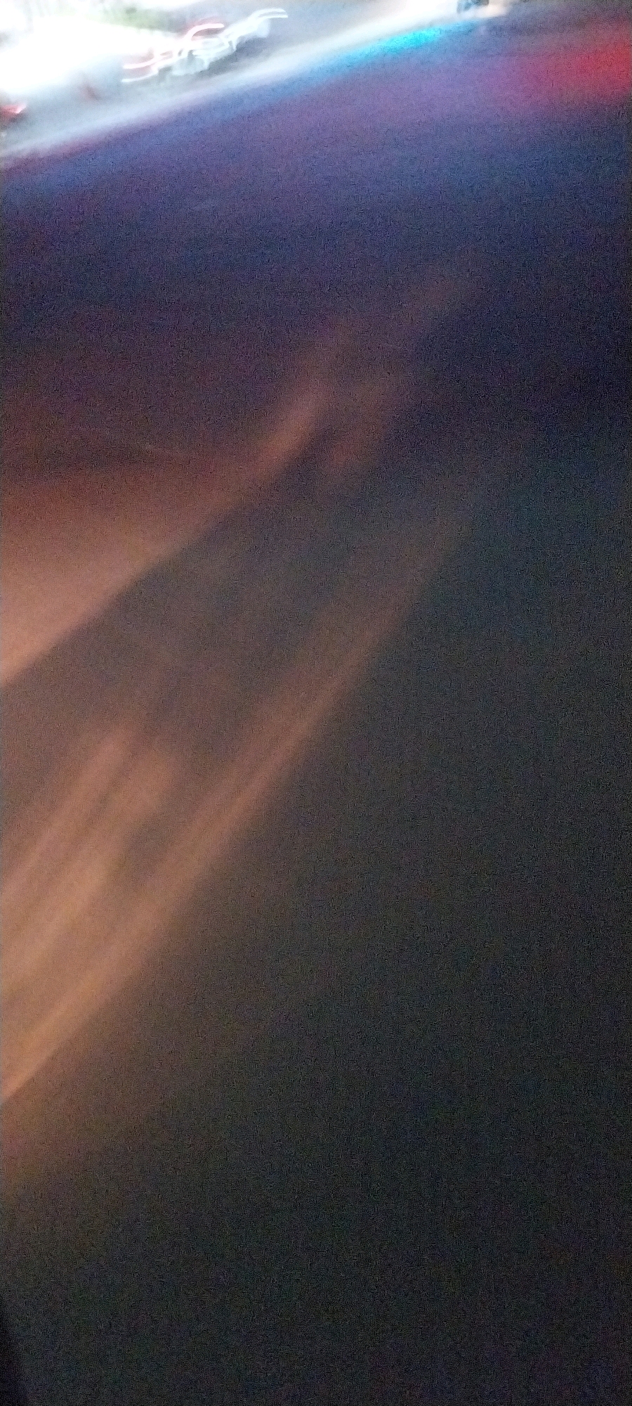 The image depicts a blurry, dark scene with streaks of light. In the upper left corner, there are hints of illuminated objects, possibly vehicles, with red and white elements and a blue light. The rest of the image is dominated by a dark, possibly nighttime, environment with diagonal streaks of orange and pink light that appear to be reflections or light beams. The specific location in Kelapa Dua, Indonesia, cannot be determined from the visual information provided. There are no clearly identifiable people, subjects, or specific actions occurring.