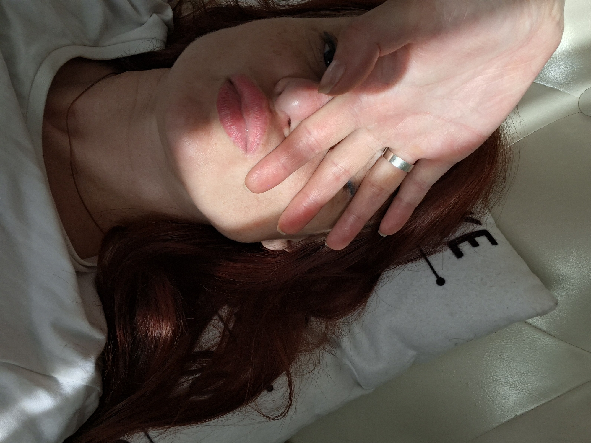 A woman with auburn hair lies on a white surface, her face partially obscured by her hand, which is adorned with a silver ring. She wears a white t-shirt and a delicate necklace. Her lips are slightly parted in a pout, and her skin is lightly freckled. A pillow with a black letter "E" and a small dot is visible behind her head. The scene is lit by sunlight casting shadows across her face. No information about the location in Myadzyel, Belarus is present in the image.