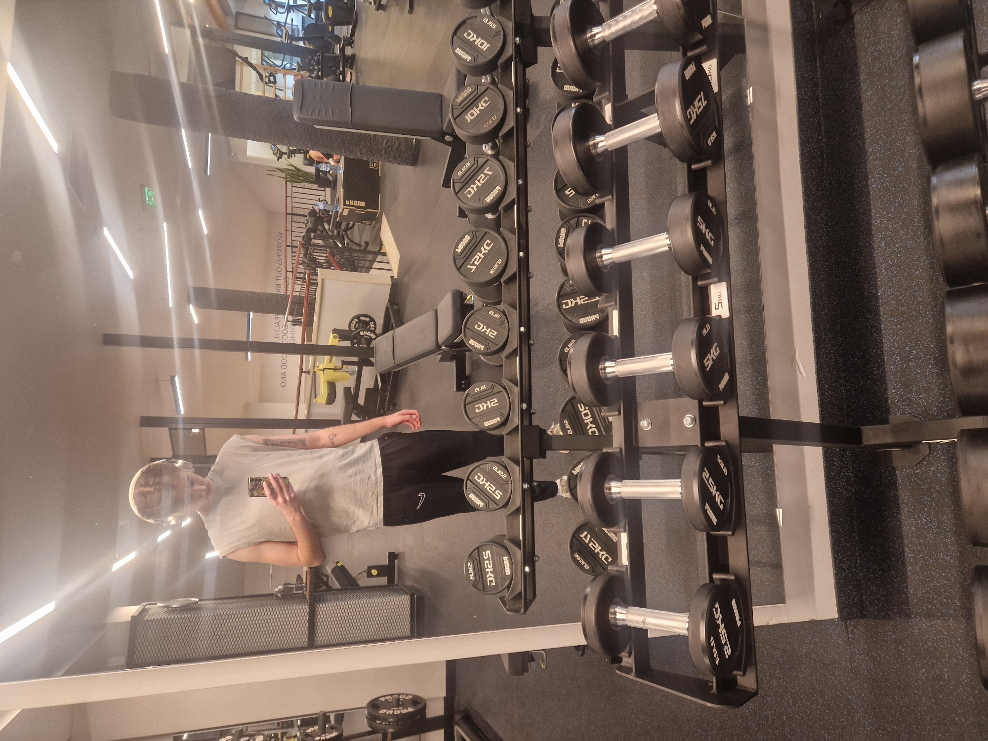 Here is a detailed description of the image:
**Scene:** An indoor gym setting. The viewpoint is a reflection in a mirror.
**People:** The reflection of a person, possibly male, can be seen in the mirror. They are wearing a t-shirt and dark athletic shorts. They also appear to be wearing headphones. The person is holding a mobile phone in their hand, possibly taking the photo.
**Objects:**
- A large rack of dumbbells of varying weights, labeled in kilograms (KG).
- Weight benches.
- Exercise machines are visible in the background, reflected in the mirror.
- A reflective mirror that makes the photograph possible.
**Environment:** The gym has light-colored walls. The overall lighting is bright and even.
**OCR:** A significant amount of text (OCR) is present in the image, mostly labels of the dumbbells "2KC", "52KC", "JOKC" and "PROUD" among others.
**In conclusion:** The image shows a person taking a photo of themselves in the mirror at a gym. There are racks of weights and weight benches in the area.