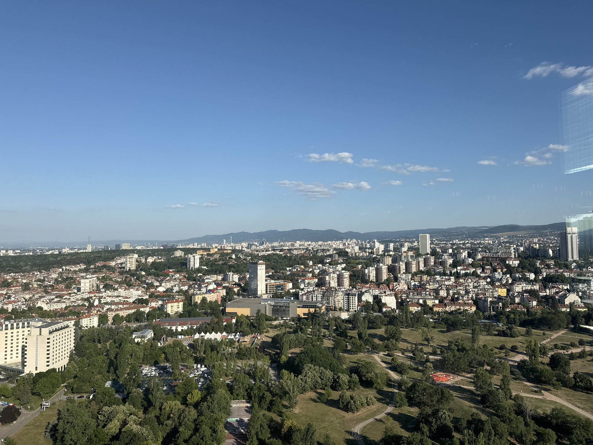 This is a wide aerial photograph of Sofia, Bulgaria, taken on a clear, sunny day. The foreground shows a park with rolling green hills, winding paths, and scattered trees. Several parked cars are visible in a parking lot on the left side of the park. Beyond the park, a dense urban landscape unfolds, characterized by a mix of low-rise buildings with red-tiled roofs and taller modern apartment buildings and office towers. The city extends towards the horizon, where a range of mountains can be seen in the distance. Several prominent structures, including a tall, slender tower and a large, modern building with a glass facade, stand out within the cityscape. The sky is a bright, clear blue with a few scattered white clouds. The reflection of windows is visible in the upper right corner of the image, suggesting the photograph was taken from a building. No people are discernible in the image.