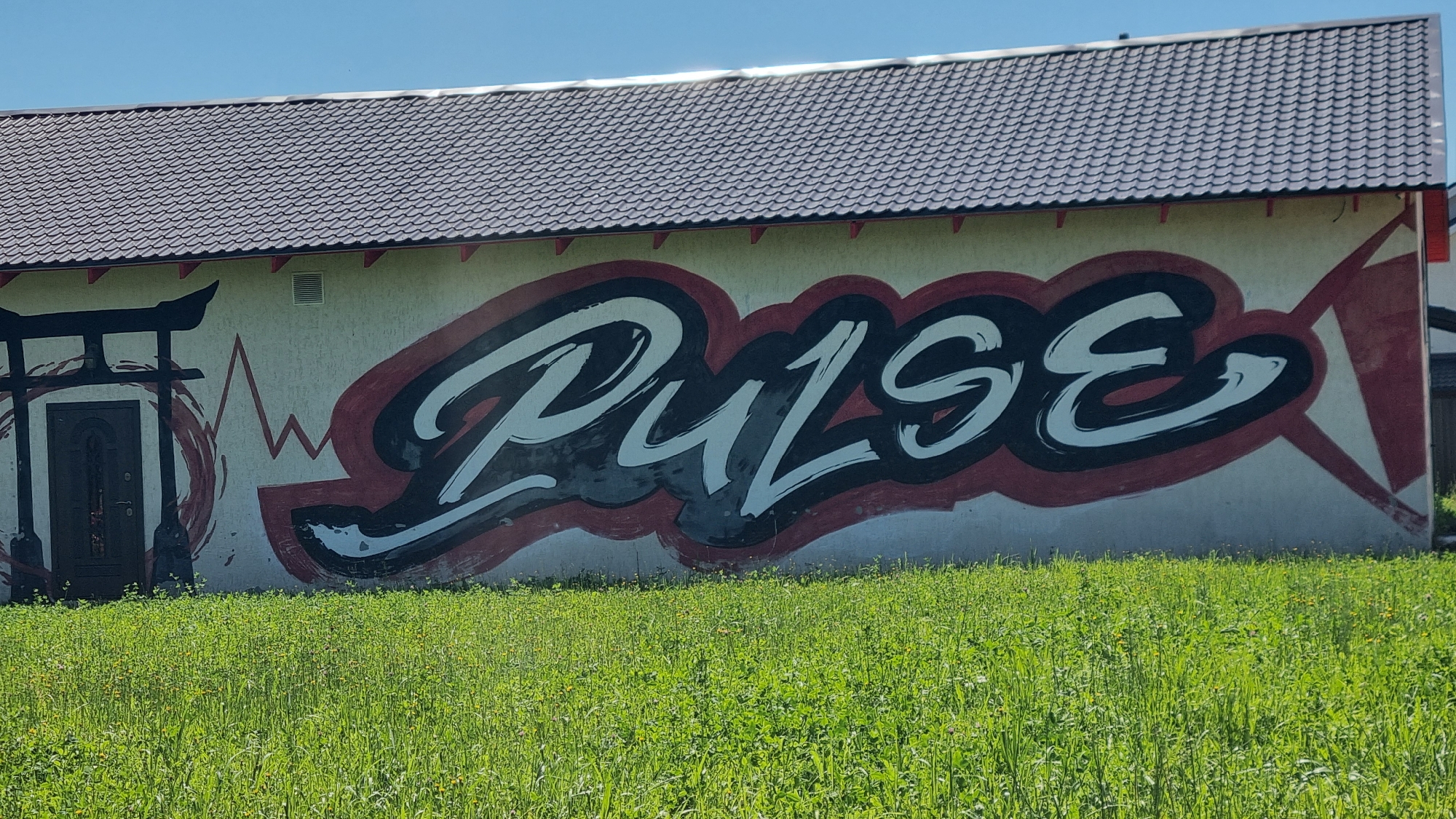 The image shows a building with graffiti on its exterior wall. The graffiti features the word "PULSE" in large, stylized white letters with black and red outlines. To the left of the graffiti, there is an illustration of a Japanese torii gate with a dark-colored door. A red, abstract waveform runs horizontally across the wall between the torii gate and the word "PULSE." The building has a dark, tiled roof and a light-colored wall. In front of the building, there is a field of green grass with scattered small yellow flowers. The sky is a clear, bright blue. This building is located in the city of Verzilovo, Russia.