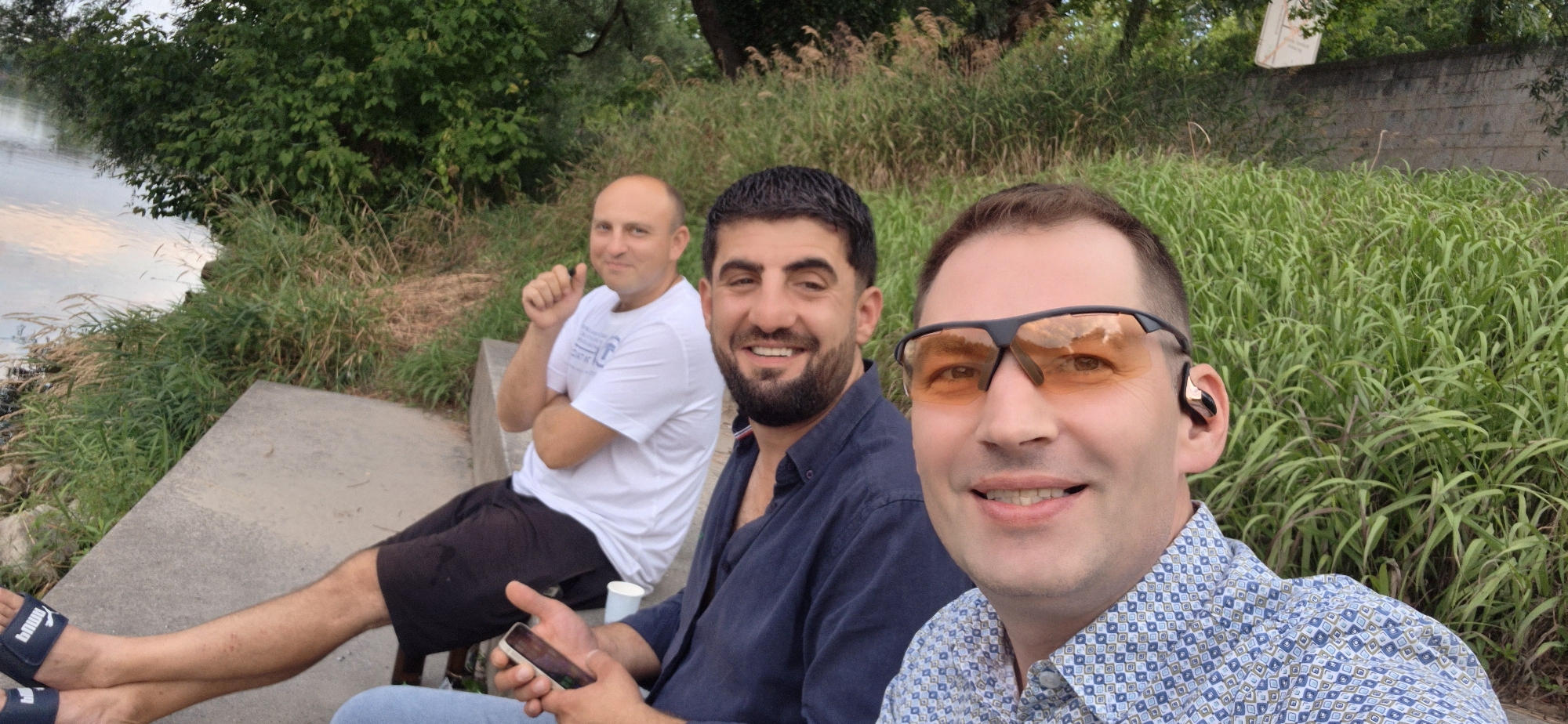 The image shows three men sitting outdoors near a body of water, possibly the Danube River in Ingolstadt, Germany. The man on the right is in the foreground, wearing a blue and white patterned shirt and orange-tinted glasses. He has short dark hair and is wearing an earbud in his right ear. He is holding a smartphone in his left hand. Next to him, to the left, is another man in a dark blue shirt, looking at the camera and smiling. He has short dark hair and a beard. The third man is seated further back on the left, wearing a white t-shirt and black shorts. He is also looking towards the camera and smiling. He has short, light-colored hair and is sitting with his arms crossed. The men are seated on a concrete ledge. Behind them, there is lush green vegetation and trees. The water is visible on the left side of the image. The overall setting suggests a casual outdoor gathering.