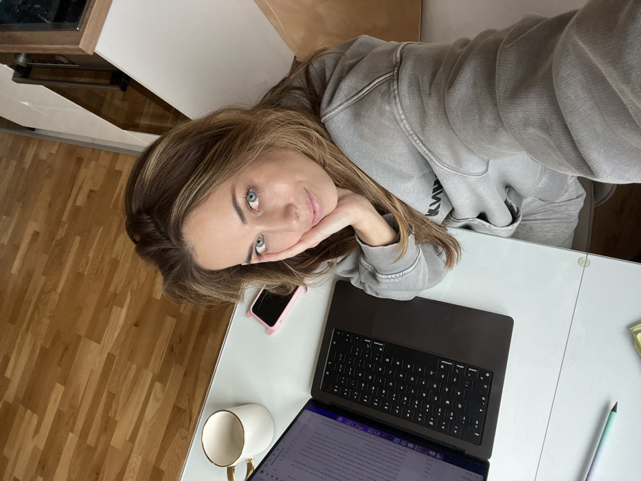 An adult female is depicted in a high-angle shot, seated at a white desk in an indoor setting within the city of Bar, Montenegro. She has light brown hair with blonde highlights and blue eyes, and is wearing a faded light gray hooded sweatshirt. Her right hand is resting on her cheek, and she is looking directly at the camera. On the white desk surface, a silver/gold MacBook Pro laptop is open, displaying text on its screen. A white mug with a gold rim and handle is positioned to the upper left of the laptop. A pink smartphone with "cat ear" protrusions is lying face down on the desk to the right of the laptop. A light blue and white pen is visible near the bottom left of the desk, along with a partially obscured small yellow-green object. The background reveals a wooden floor, white architectural elements, and reflective surfaces.