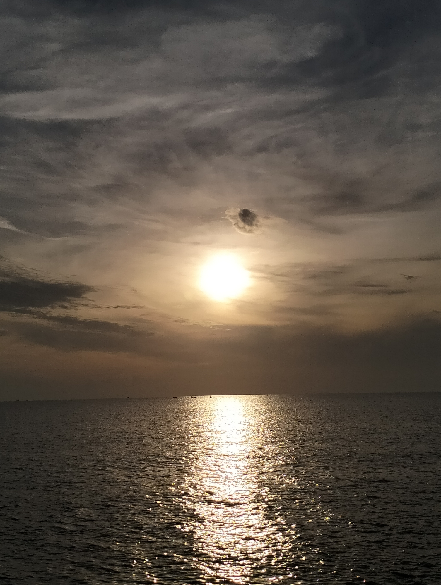The image captures a wide expanse of the ocean, with the sun partially obscured by clouds, casting a bright reflection on the water's surface. The sky is filled with a mix of dark and light clouds, creating a dramatic atmospheric effect. The horizon line is clear, separating the sea from the sky. Small boats are visible in the distance on the water. The scene is likely a coastal area in Lhokseumawe, Indonesia, known for its beaches and maritime activities.
