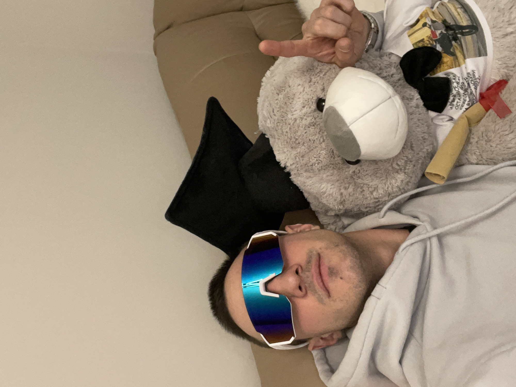 The image shows a man with dark hair wearing mirrored sunglasses and a light grey hoodie. He is lying on a couch with a grey teddy bear. The teddy bear is wearing a white t-shirt, a black bow, and has a rolled-up object with a red piece attached to it. The man is holding his hand up near the teddy bear's head, with his index finger extended. A black pillow is also visible. The background shows a beige couch and a light-colored wall. The location in Plovdiv, Bulgaria, cannot be determined from the visual information provided.