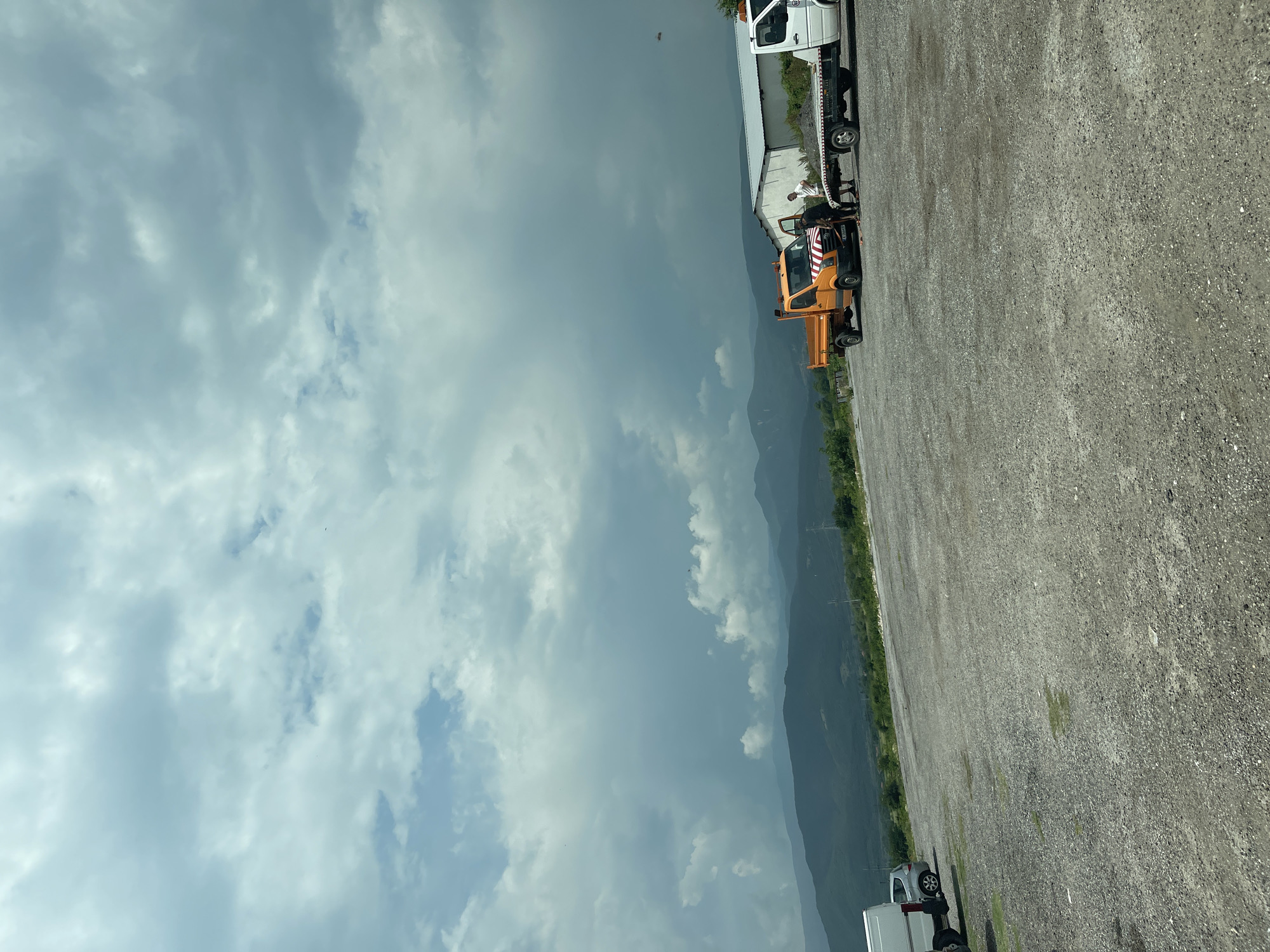 This image depicts an outdoor, unpaved gravel lot located in Karnare, Bulgaria, under an overcast sky. The horizon reveals multiple layers of mountains extending into the distance, appearing progressively lighter in tone due to atmospheric perspective. In the mid-ground, a simple, white, single-story building with a light-colored roof is situated on the right side. Utility poles and power lines are visible in the landscape between the building and the mountains.
The gravel lot itself shows sparse patches of green vegetation. On the far left, a portion of a white panel van is parked, with a silver hatchback visible behind it. Closer to the center, an orange dump truck is parked with its driver's side door open. This truck features a distinctive red and white striped reflective decal across its front bumper/grille. Adjacent to the orange truck, a white flatbed tow truck is positioned, pulling a trailer that is loaded with a dark, granular material, likely gravel or soil.
Two male individuals are present in the scene. One man, dressed in a dark shirt and light trousers, is seen bending down near the open door of the orange dump truck, appearing to be engaged in an activity involving the vehicle. The second man, wearing a white t-shirt and dark trousers, stands upright between the orange truck and the white flatbed truck, observing the activity.