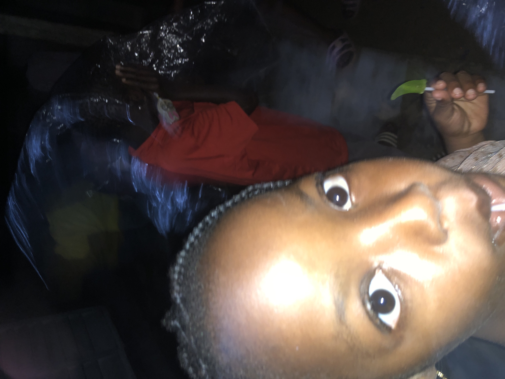 A close-up, low-light, first-person perspective shot captures a young person's face in the foreground, with a blurry background. The individual's dark skin and braided hair are visible, with one eye and part of the nose and mouth in sharp focus. They are holding a green, leaf-shaped object on a white stick, possibly candy or a novelty item, in their right hand. In the background, a person in a bright red garment is partially visible, their face obscured. Other figures and objects are indistinct due to motion blur and low lighting, suggesting a busy or dynamic environment. The scene appears to be indoors or in a dimly lit outdoor setting, potentially a gathering or event. The overall image quality is poor due to low resolution, poor lighting, and significant motion blur.
