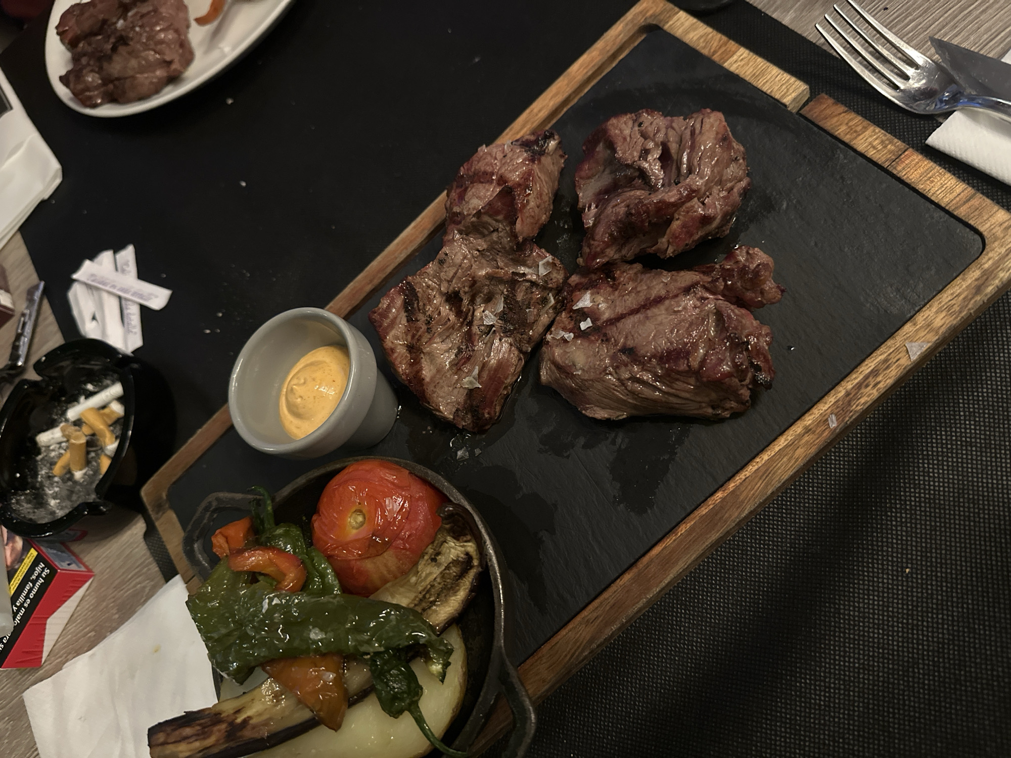 The image shows a meal on a table in what appears to be a restaurant setting. The main feature is a wooden serving board with a dark, possibly slate, surface. Arranged on the board are pieces of cooked meat, a small dish of yellow sauce, and a metal skillet containing roasted vegetables including a tomato, eggplant, potatoes, and green peppers. To the left of the serving board is an ashtray full of cigarette butts and a packet of cigarettes. Utensils (fork and knife) and a folded napkin are visible to the right of the serving board. A plate with more meat is partially visible in the upper-left corner. The scene is in Sant Feliu De Guíxols, Spain.