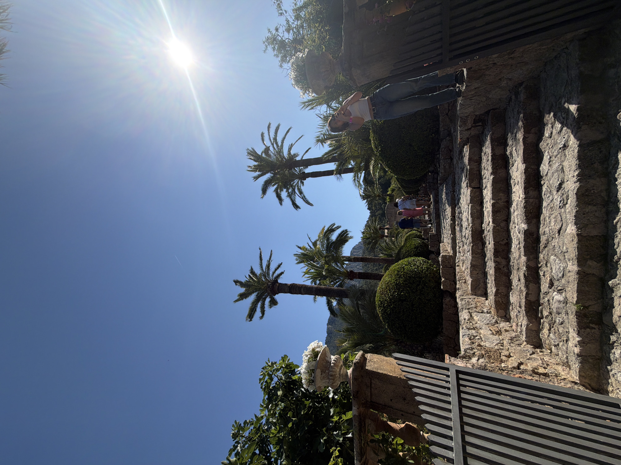 The image shows an outdoor scene in Bunyola, Spain, likely at a garden or park. A stone staircase ascends alongside a wall, with various plants and trees present, including palm trees and round bushes. Sunlight is visible in the upper left of the frame. A woman is standing near the top of the stairs, and other people are visible in the middle distance. A metal gate is in the foreground.