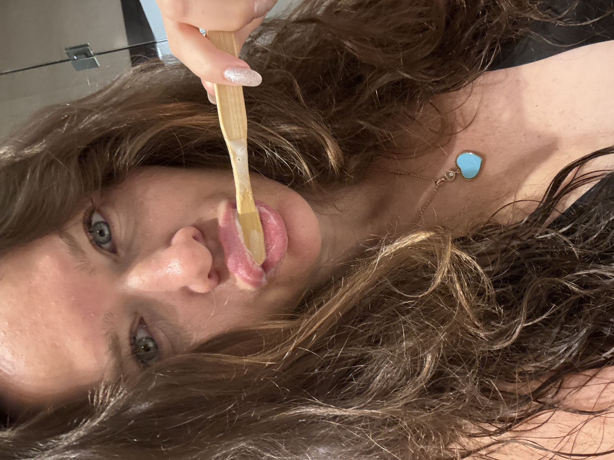 The image shows a close-up of a woman's face as she brushes her teeth. She has long, brown, wavy hair and light blue eyes. She is wearing a black top and a necklace with a blue heart-shaped pendant. The toothbrush is made of bamboo and has white toothpaste on it. The scene appears to be in a bathroom, possibly in Dubai, United Arab Emirates, given the context provided. The lighting is artificial and the background is blurred, suggesting an indoor environment.