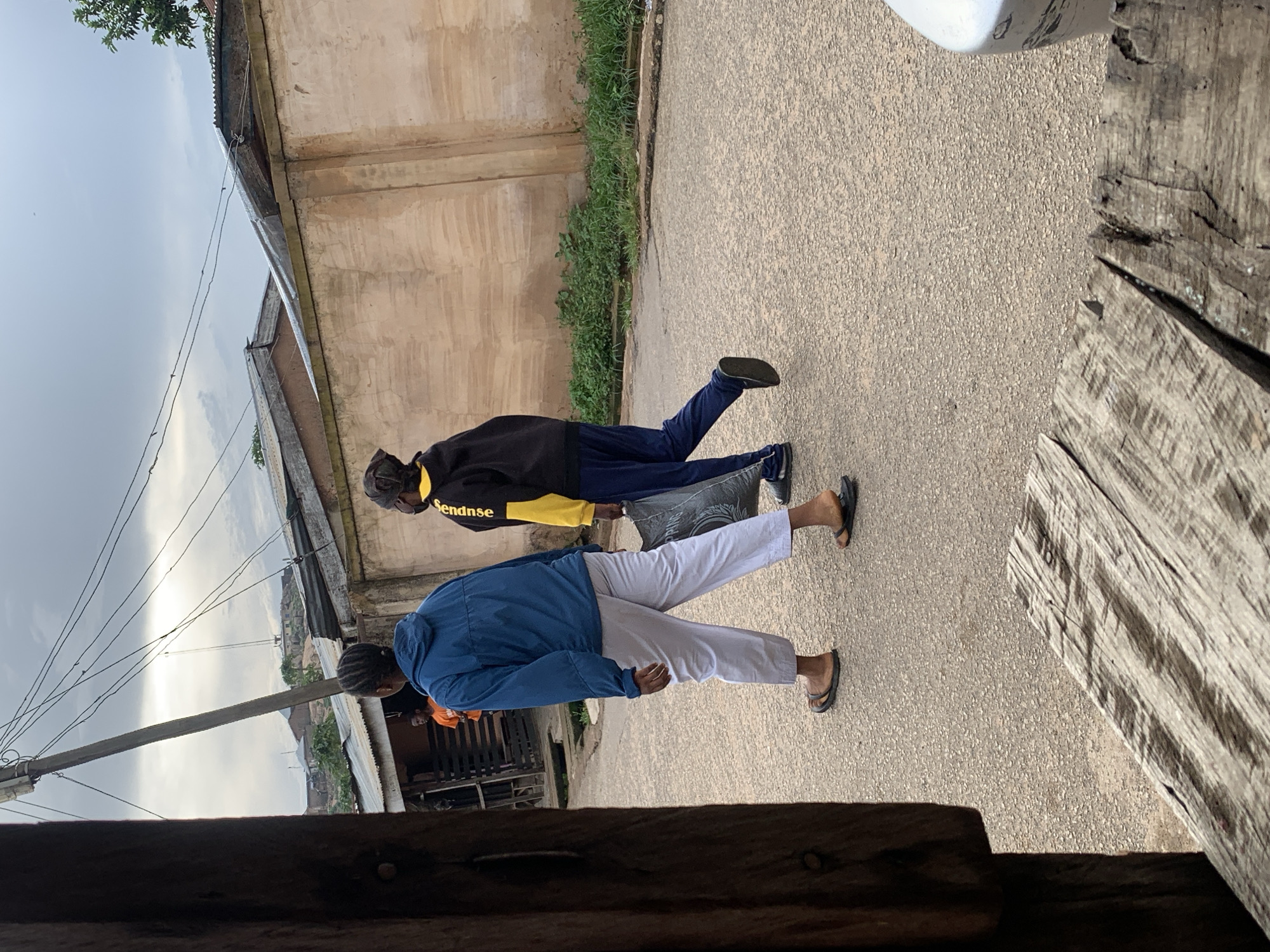 Two individuals walk away from the camera on a paved road in Jos, Nigeria. The person on the left wears a blue hooded jacket and light gray pants, with dark sandals. The person on the right wears a black and yellow hooded jacket and dark blue pants, carrying a large black bag. In the background, a concrete wall, corrugated metal roofs, and vegetation are visible under an overcast sky. A wooden structure is in the foreground on the left, and a portion of a white vehicle is on the right. A utility pole with numerous wires stands to the left of the individuals.