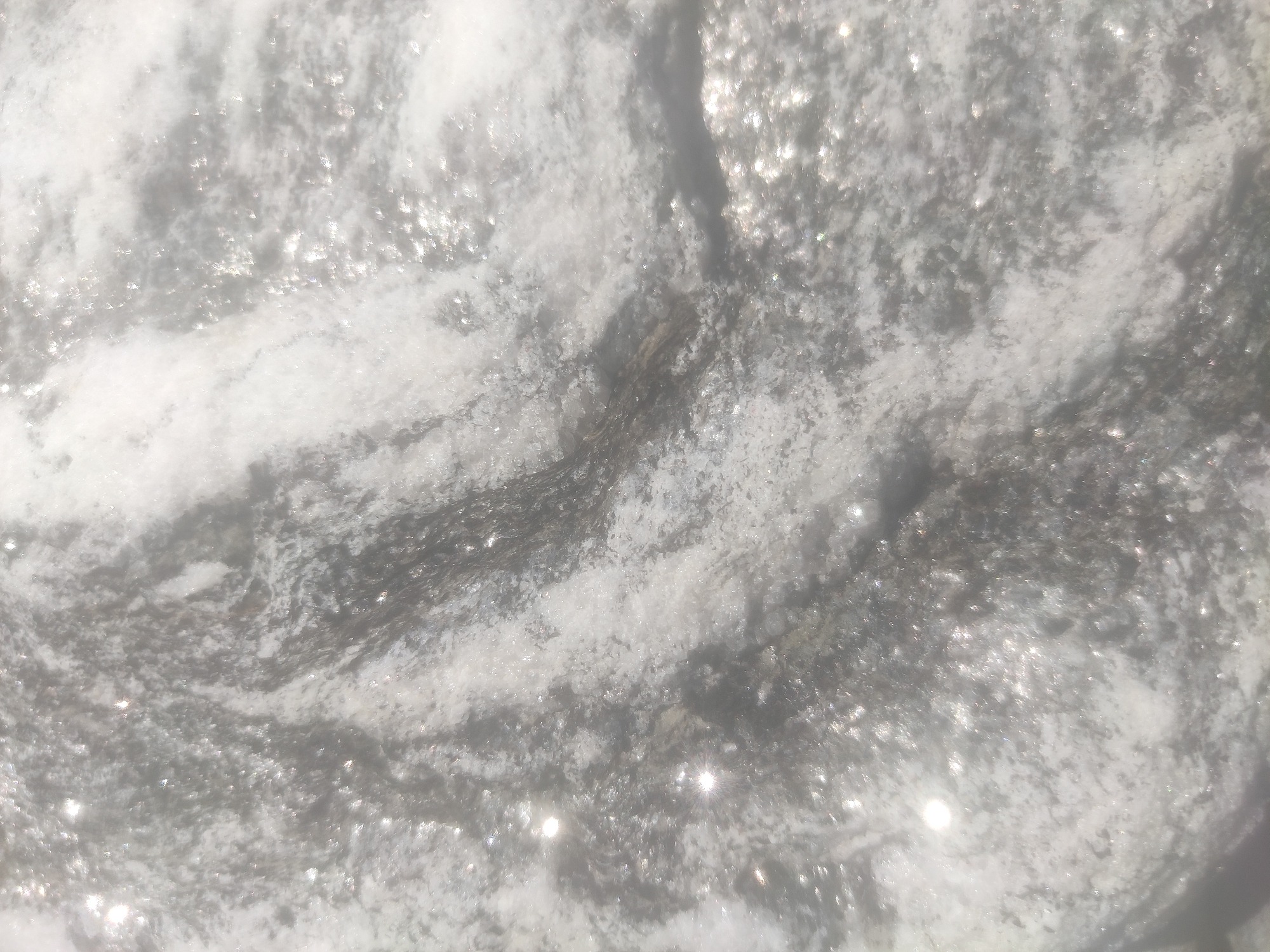 The image shows a close-up view of a textured surface, possibly a rock or stone, with swirling patterns of white and gray. There are small, sparkling highlights scattered across the surface, suggesting the presence of light reflecting off small particles or crystals. The texture appears rough and uneven. No people or specific objects are discernible, nor are any actions or interactions depicted. The overall impression is of a natural, mineral surface. The location in Katrain, India, is not visually represented in the image itself.