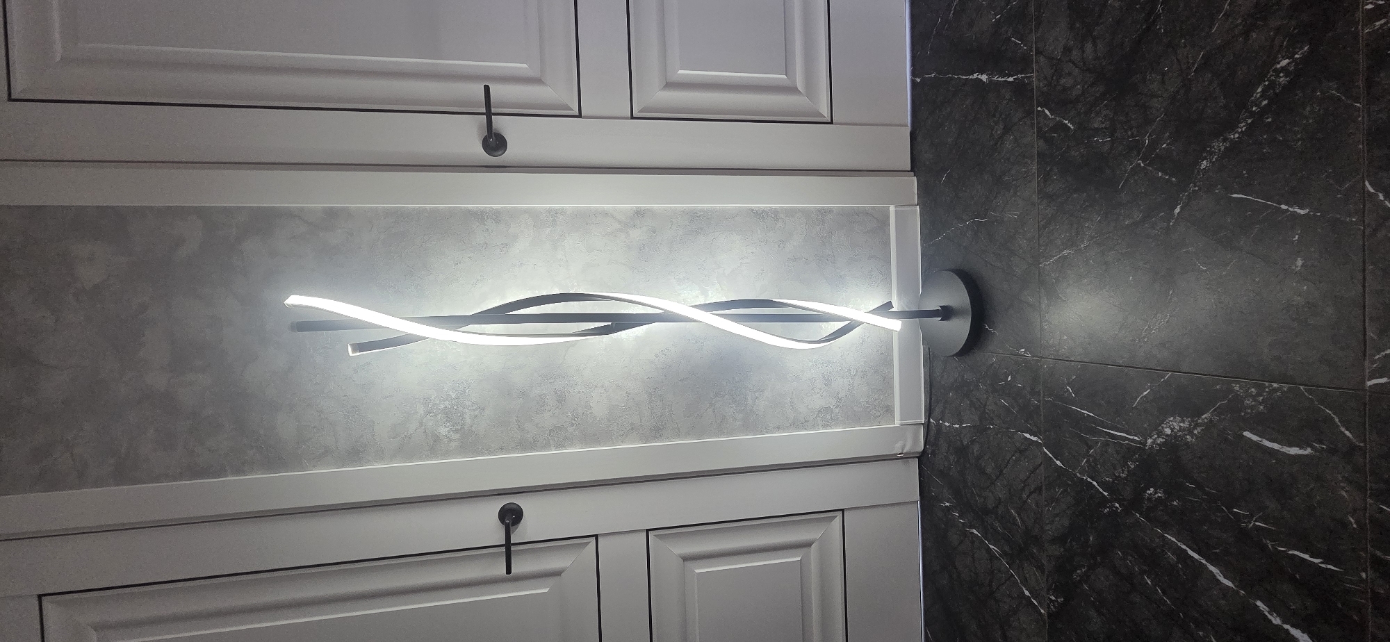 The image shows a modern LED floor lamp with a black base and a twisted design featuring a white light source. The lamp is positioned in the center of a hallway, between two white doors with black handles. The wall behind the lamp has a textured, gray finish. The floor is tiled with a dark marble pattern. The scene appears to be indoors, likely in a residential or commercial setting. No specific location in the city of Markova, Russia, can be identified from the image.