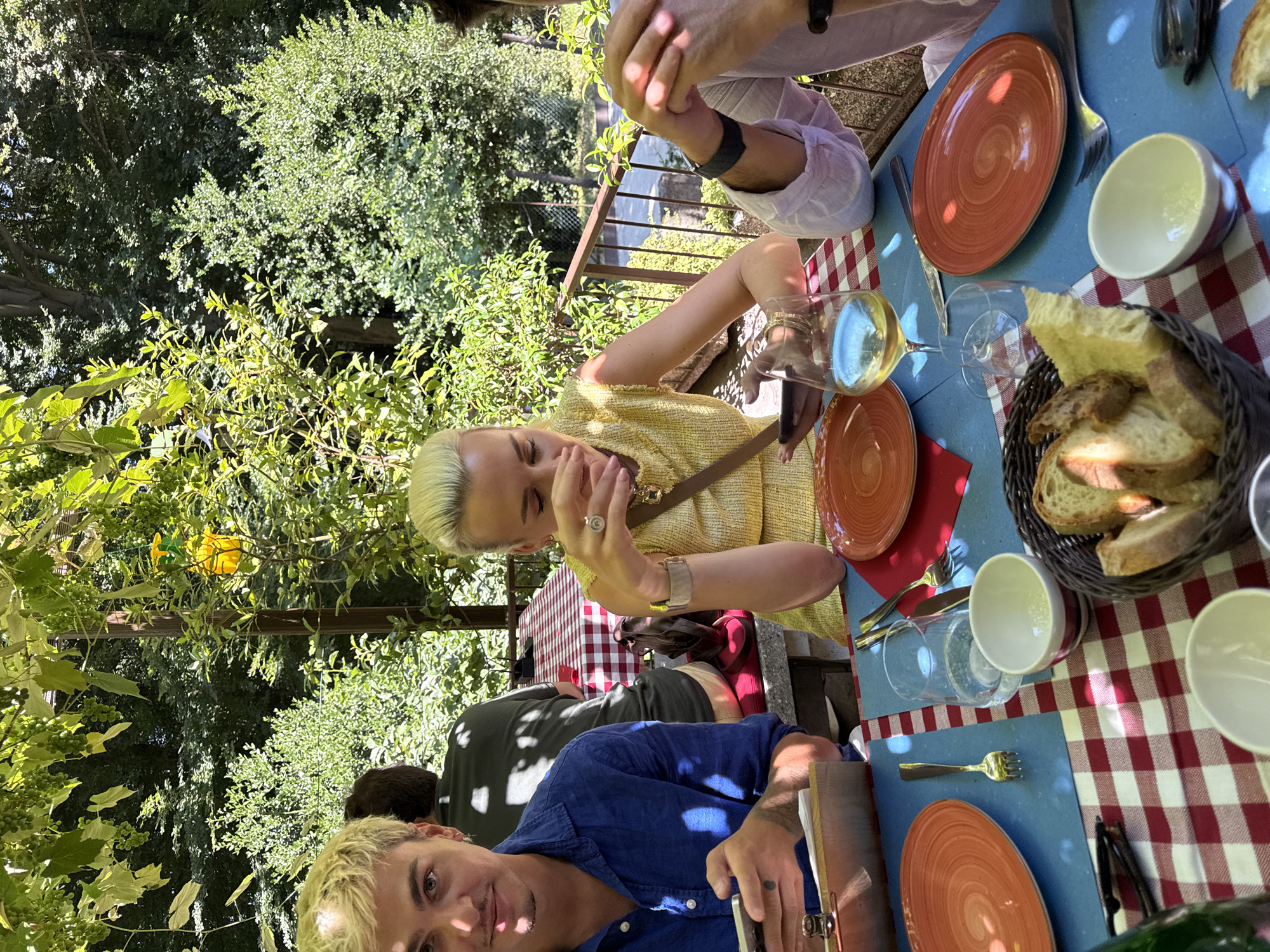 A group of people are seated at an outdoor table with a red and white checkered tablecloth. The table is set with orange plates, white bowls, and cutlery. In the foreground, a basket of bread is visible. A person with blonde hair, wearing a yellow top, is seated in the middle of the frame, looking down at their phone. To the left, a person with blonde hair and a blue shirt is also looking at their phone. In the background, trees and foliage create a lush, green environment, suggesting a location in a natural setting. There is also a yellow object hanging from a tree. The scene appears to be a casual meal outdoors, possibly at a restaurant in Agno, Switzerland, given the presence of the checkered tablecloth and the outdoor dining setup.