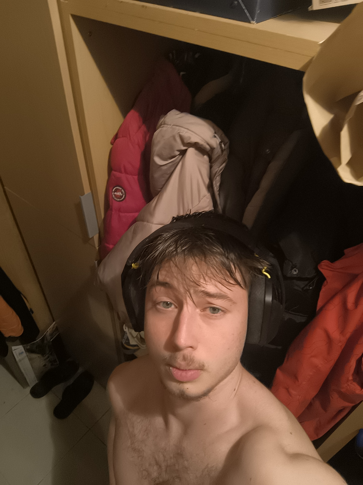 A shirtless young man with fair skin, light brown hair, and a mustache is wearing black over-ear headphones with yellow accents. He is positioned in front of an open closet or wardrobe filled with jackets in various colors, including pink, beige, black, and red. On the floor to the left, there are two black shoes and a plastic bag. The scene appears to be indoors. There is no explicit information to place the location in the city of Sofia, Bulgaria.