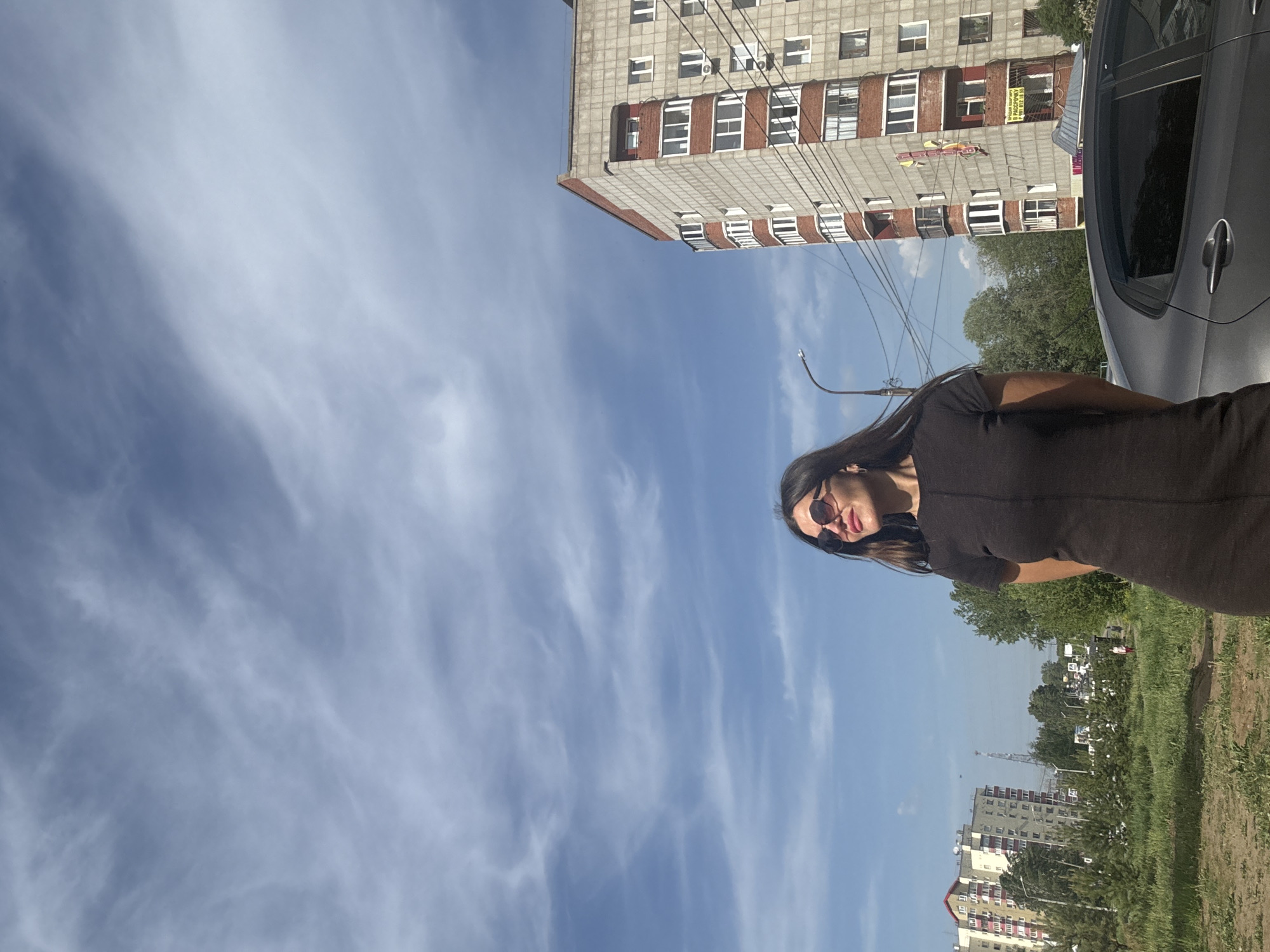 The image captures a woman wearing a dark dress and sunglasses, standing outdoors in Chaikovsky, Russia. In the background, there are residential buildings, trees, and a blue sky with scattered clouds. A portion of a gray car is visible to the right. The scene appears to be a street or sidewalk in the city.
