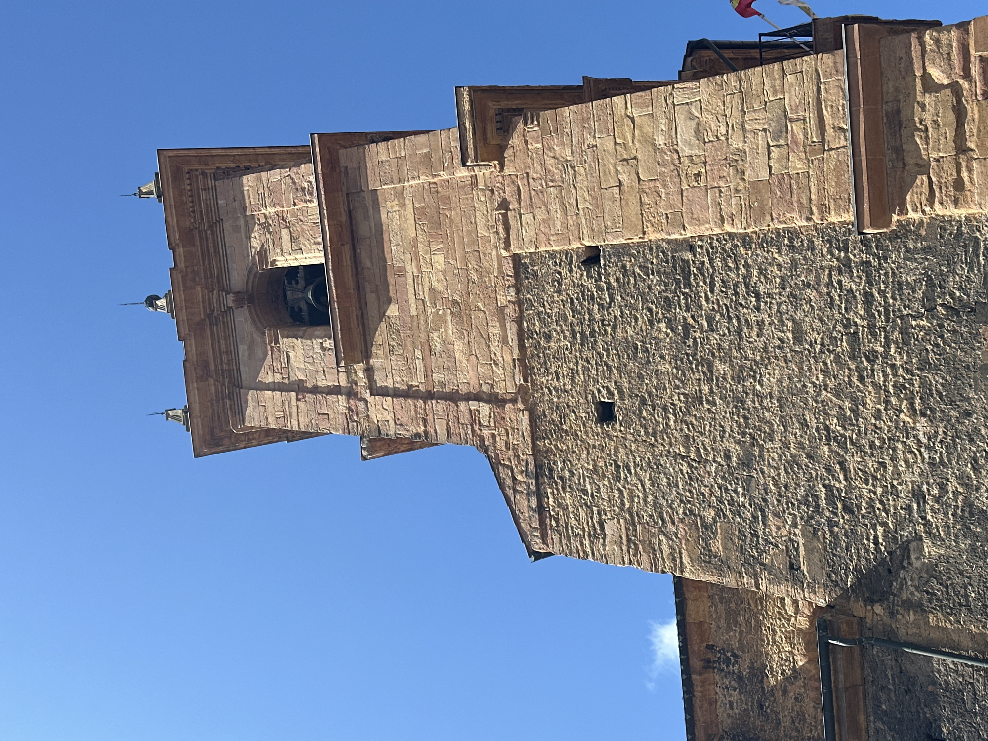 The image captures a close-up, low-angle view of a stone bell tower, likely part of a church or cathedral, against a clear, bright blue sky. The tower is constructed from rough-hewn, reddish-brown stone blocks of varying sizes, giving it a textured and aged appearance. The stonework shows signs of weathering and has a rustic, somewhat uneven finish.
The tower features architectural details such as cornices and decorative elements. At the top, a belfry section is visible, containing a dark-colored bell. Several spires or decorative finials surmount the uppermost part of the tower. Sunlight illuminates the stone, casting shadows that highlight the architectural features and the rough texture of the masonry.
The lower portion of the tower visible in the image is of a more solid, less decorated design, with small, dark openings that might be windows or ventilation shafts. Part of an adjacent structure, also made of stone, extends from the left side of the frame, with a dark roofline and what appears to be a downspout. A black metal railing is visible on the right side of the tower, suggesting a balcony or observation point. A small portion of a flag with red and yellow colors is also visible on the far right.
The overall impression is one of historical architecture bathed in strong daylight. The image does not contain any people, and no specific actions or interactions are depicted. The environment is solely characterized by the clear blue sky and the imposing stone structure. The context of Oviedo, Spain, would suggest this is a historical religious building, common in the city's architectural landscape.