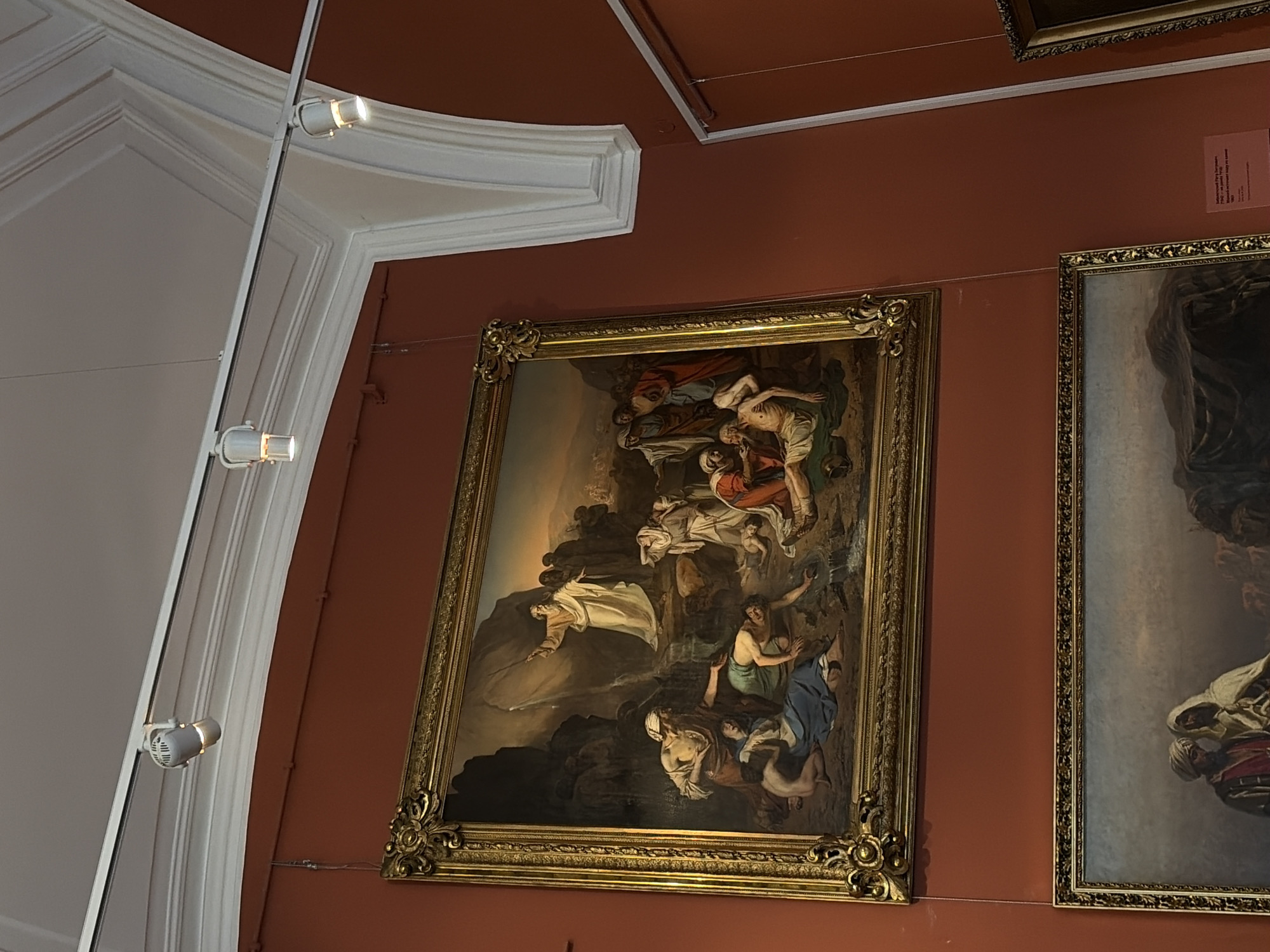 The media file shows two large, ornate, framed paintings displayed on a burnt orange wall. The painting on the left is the primary focus, depicting a dramatic scene with multiple figures. The scene appears to be religious or mythological, with figures in flowing robes set against a dark, rocky landscape and a dramatic sky. One prominent figure, draped in white, is reaching out with an expression of anguish or entreaty. Other figures are shown in various states of distress or contemplation, some tending to or supporting others. The style of painting suggests a historical or classical period.
The painting on the right is partially visible and appears to be in a similar style, featuring figures in historical attire against a muted background. A small, pink label with text in Cyrillic script is attached to the wall to the right of the central painting. The lighting in the room consists of track lighting with white cylindrical spotlights, two of which are clearly visible on a white architectural element that frames the top left of the image. This architectural element has classical moldings, suggesting the location is a museum or gallery, likely a grand one given the ornate frames and the wall treatment. The overall impression is one of a formal exhibition space, potentially in Saint Petersburg, Russia, as indicated by the location context. No specific time of day or weather conditions are discernible from the artwork itself. The emotions conveyed are predominantly somber and intense.