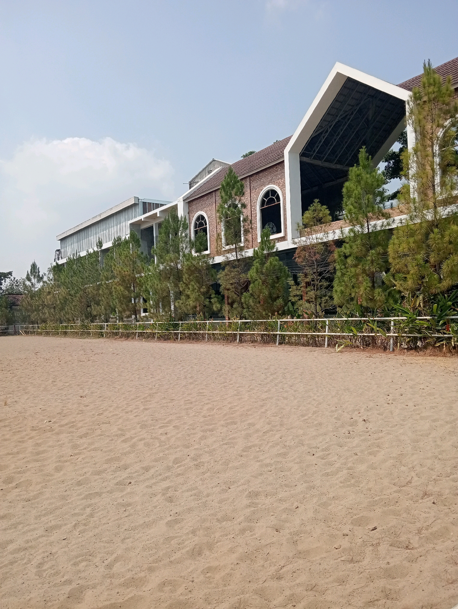 A long, sandy expanse occupies the foreground of this image, its surface textured by subtle undulations and scattered debris. A low, white fence runs horizontally across the middle ground, demarcating the edge of the sandy area. Beyond the fence, a line of evergreen trees, appearing to be pine or fir, stretches across the scene. Behind the trees, a building complex rises, characterized by a mix of architectural styles. On the right, a section of the building features a prominent, steeply pitched white roof structure with exposed beams, set against a backdrop of brickwork and arched windows. To the left of this section, a longer, more modern wing of the building is visible, with a facade dominated by horizontal window elements and metallic sheathing. The overall impression is of a resort or commercial establishment situated in a semi-urban or coastal environment, likely in Pringsewu, Indonesia, given the sandy terrain and lush greenery. The sky above is a pale, hazy blue with a single, wispy cloud. No people are visible in the frame.