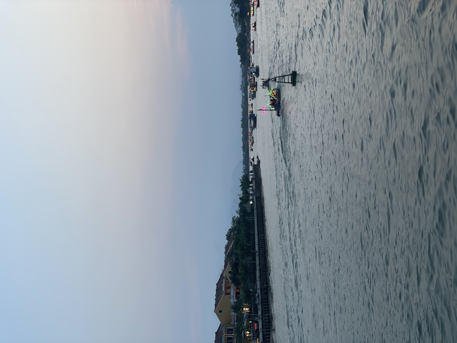 An evening scene on the Thu Bồn River in Hội An Ward, Vietnam, is depicted. The sky, occupying the upper two-thirds of the image, displays a gradient from light blue-grey at the zenith to a darker purple-grey near the horizon, with a subtle pink tint on the far right. The lower half of the frame consists of the river's surface, characterized by dark, choppy, grey-blue water with visible ripples. Numerous boats are present on the water, moving generally away from the viewer and towards the left mid-distance. Many of these vessels are illuminated with bright, multi-colored lights (red, green, blue, yellow), suggesting a festive or tourist activity. A prominent green, conical navigational buoy with a white top is positioned in the mid-right foreground. Along the left bank, a series of traditional-style buildings with yellow-ochre facades and reddish-brown tiled roofs are visible, interspersed with green foliage. An elevated dark-colored promenade or pier runs along the riverbank in front of these structures, with artificial streetlights and indistinct figures indicating pedestrian presence. Hazy, distant mountains are faintly visible on the horizon behind the buildings.