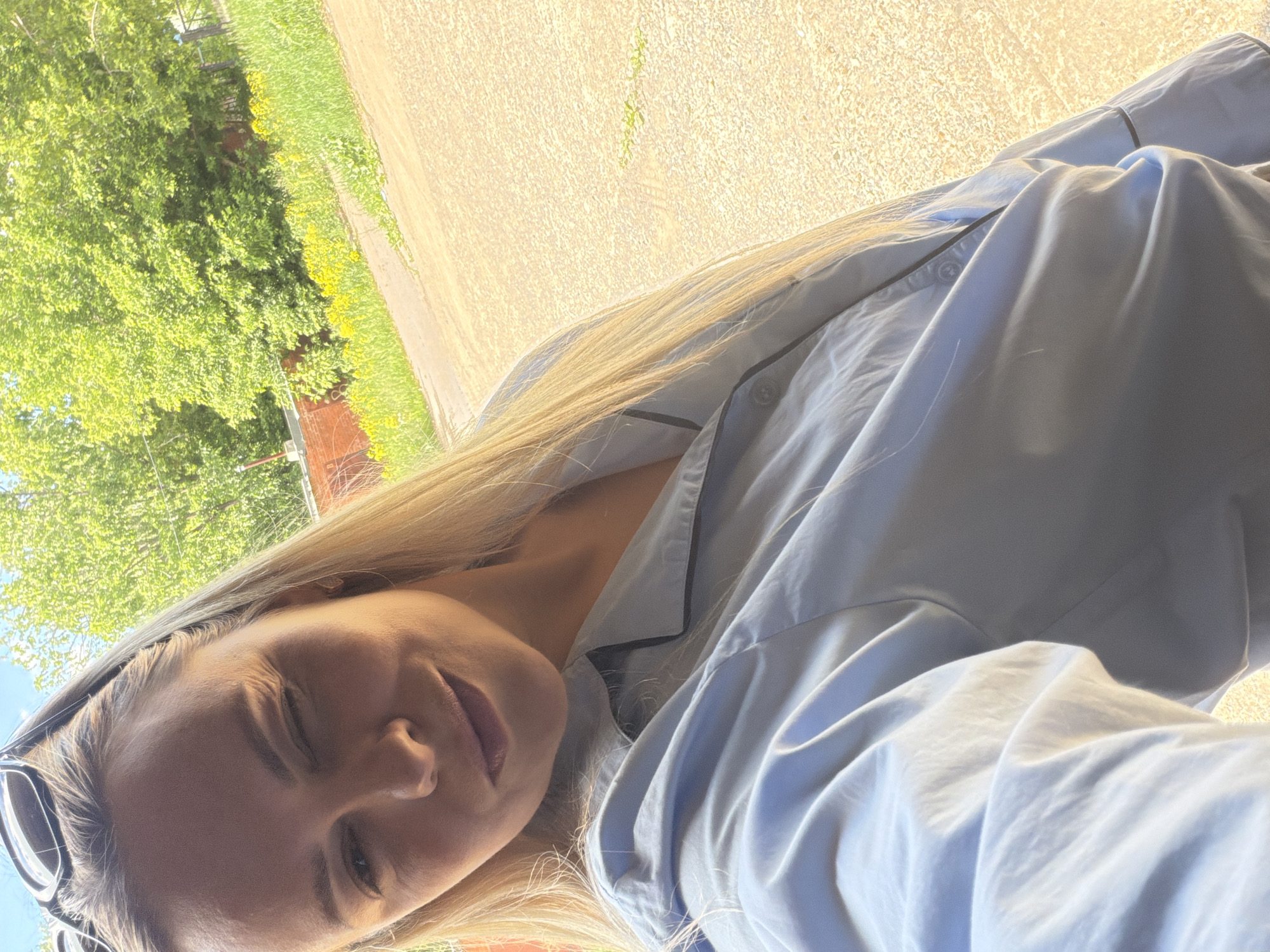 This media file features a candid, outdoor self-portrait of a young woman, likely taken in Votkinsk, Russia, as indicated by the location context. The image is bright and appears to be taken in strong daylight.
The main subject is a woman with long, straight blonde hair, which cascades over her shoulder and chest. She is looking towards the camera with a gentle smile or smirk, her eyes partially closed, suggesting either squinting from the sun or a relaxed, playful expression. She is wearing a light blue button-up top, possibly a pajama shirt or a casual blouse, with noticeable dark piping along the collar and front placket. Sunglasses are perched on top of her head, indicating she was outdoors in sunny conditions.
The background shows a lush, green environment. To the left, there are dense green trees and foliage, with patches of small yellow flowers or weeds visible amongst the greenery. To the right and behind her, a wide, textured expanse of light brown or sandy-colored ground is visible, possibly a dirt road or a paved path. The overall impression is one of a bright, sunny day in a natural or semi-natural outdoor setting. No visible text is present in the image.