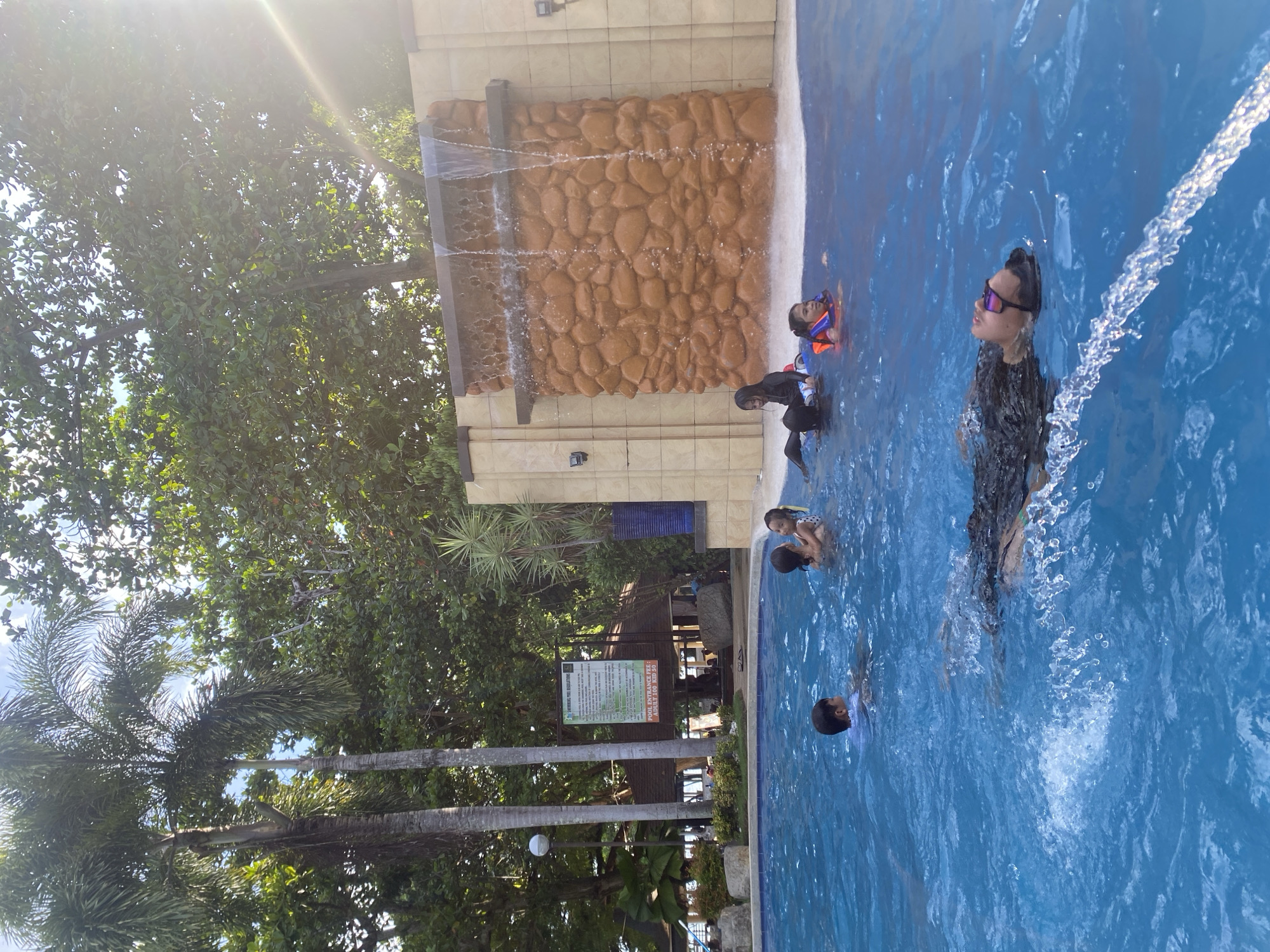 This outdoor scene in Banaybanay, Philippines, shows a swimming pool with several people. A man wearing goggles is standing in the water, near a waterfall feature built into the side of the pool. Nearby, a child wears a life vest. Others are swimming in the pool, which is bordered by trees and some built structures.