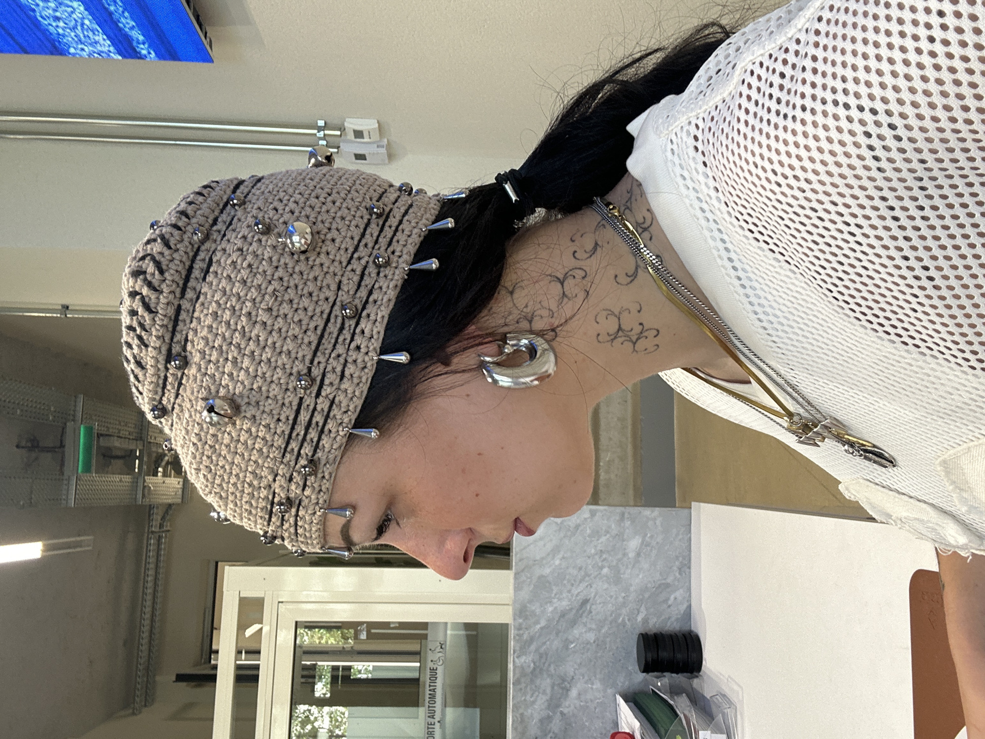 A woman wearing a studded crochet hat and a white, mesh-like top is indoors, possibly in a commercial or industrial space in Paris, France. She is positioned to the right of the frame, seen in a profile view from the shoulders up. The hat is beige with rows of silver studs and spikes. The woman has dark hair, pulled back in a ponytail, visible behind her ear, and a silver hoop earring. Tattoos are visible on her neck. She wears multiple silver necklaces. The background shows exposed ceiling pipes and a window/door with French text, "PORTE AUTOMATIQUE". A marble-patterned surface and white objects are on the right side of the frame, suggesting a table or counter.