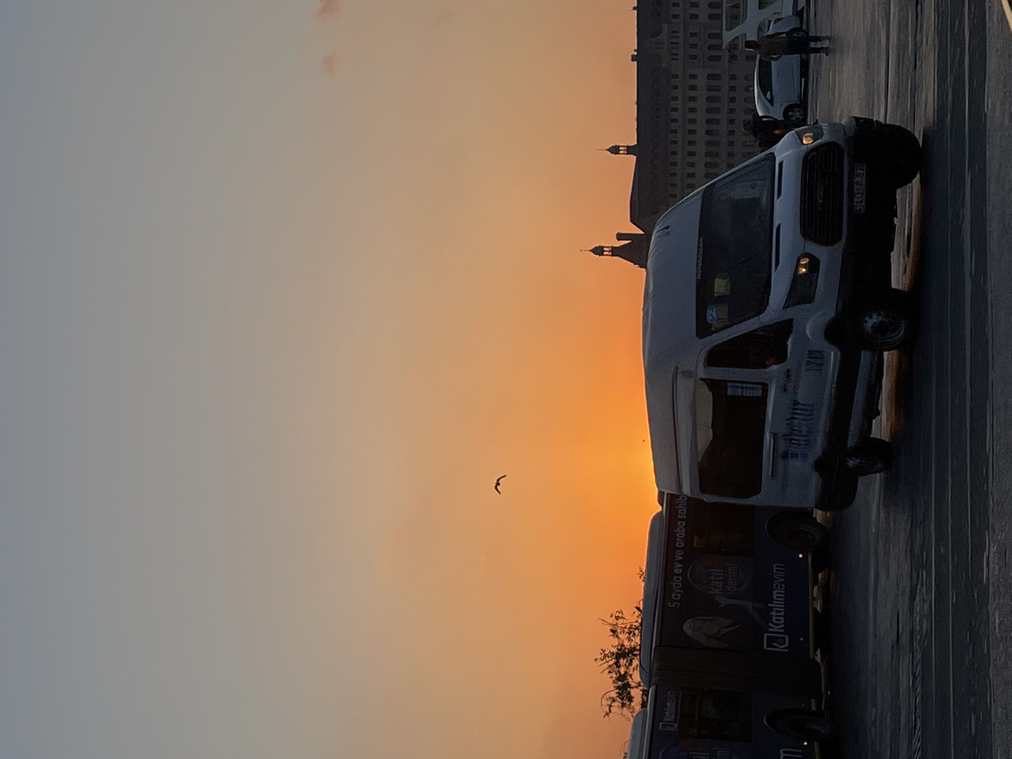 At dusk in Kadıköy, Turkey, a white van is parked in the foreground, its headlights on. Behind it, a dark-colored bus with advertisements on its side is also visible. In the distance, the silhouette of a large building with architectural details, possibly a historical structure or a government building, stands against the vibrant orange and yellow hues of the setting sun. A solitary bird is captured in flight in the sky, adding a dynamic element to the serene twilight scene. The ground appears to be a paved area, likely a public square or street.