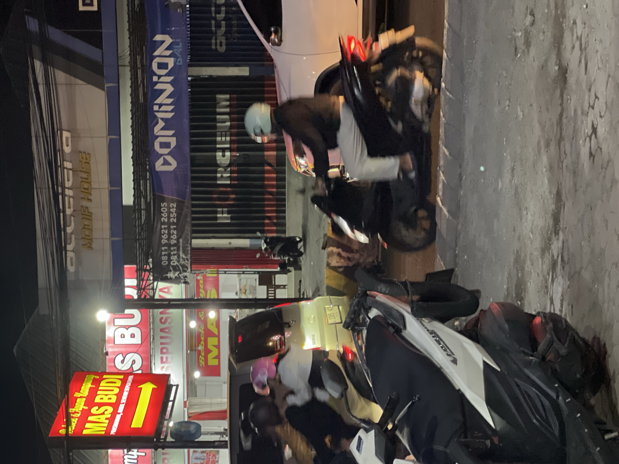 This is a nighttime street scene in Denpasar, Indonesia, likely near a commercial area, as indicated by multiple signs. In the foreground, two motorcycles are parked. The left motorcycle is white and black, and the right motorcycle is black with a rider wearing a light blue helmet and white pants, riding away from the camera. In the middle ground, a light-colored van with the name "SERENA" visible on the back is stopped, with two individuals, one wearing a pink helmet, interacting near it. Behind the van, several businesses are visible. A prominent red sign with yellow lettering reads "MAS BUDI" and features a yellow arrow pointing to the right. Another red sign behind it also says "MAS BUDI." Further back, a dark banner with white lettering states "DOMINION BALI" and the text "FORCEUM" is visible on a dark, ribbed structure. The environment appears to be a paved road with some debris, and overhead wires are visible against the dark sky. The lighting is artificial, coming from streetlights and signs, creating shadows and highlights.