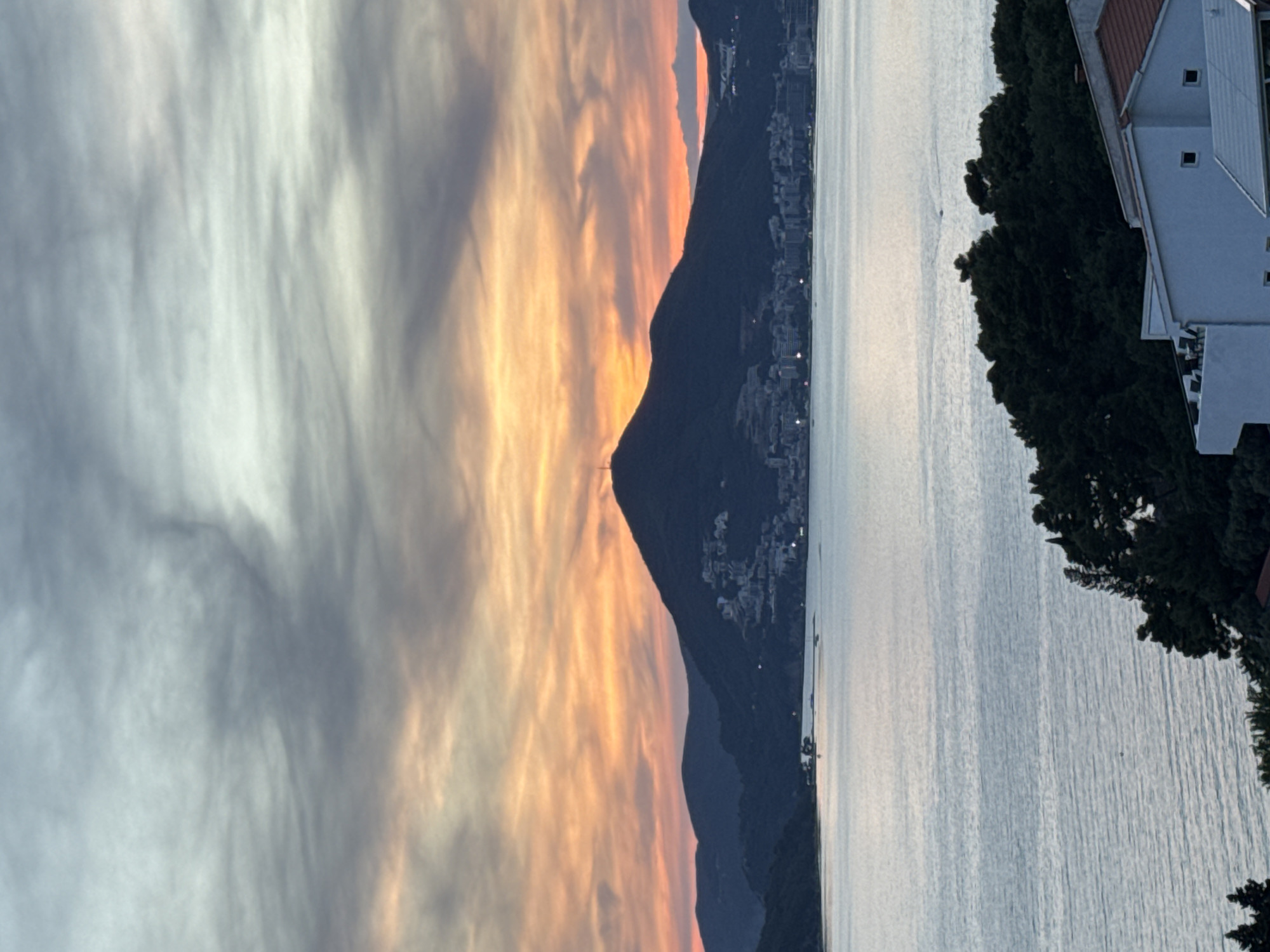 The image shows a coastal scene with a dramatic sky at sunrise or sunset. To the left, clouds display shades of gray, orange, and pink. In the center, a dark, mountainous island rises from the water. Buildings are visible on the island's slope, suggesting a populated area. The right side of the image shows a body of water with a calm surface and a building with a white facade and multiple windows. The building is partially obscured by dark green foliage. The location is identified as Sveti Stefan, Montenegro.