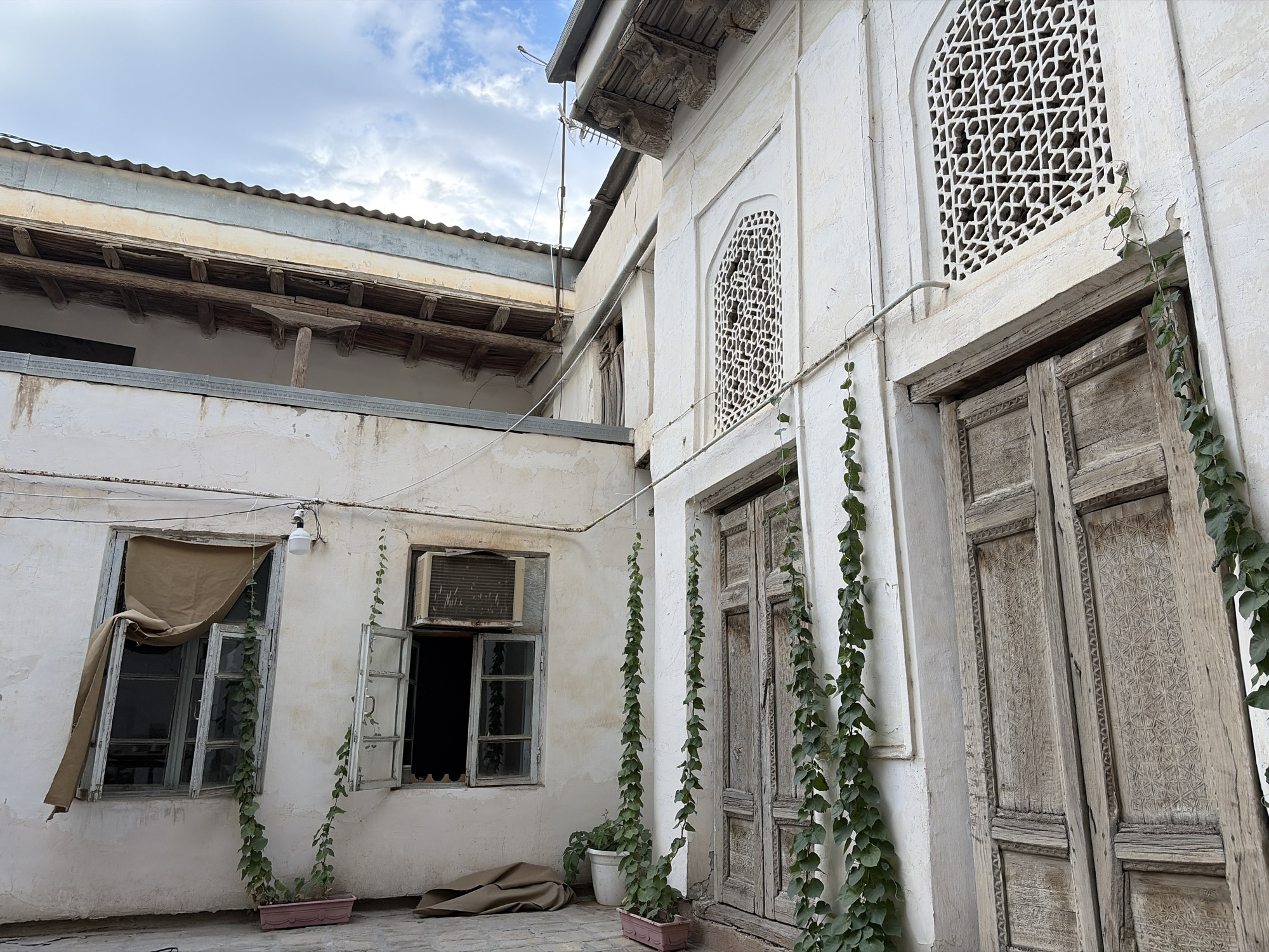 The image depicts a courtyard in Bukhara, Uzbekistan, featuring weathered traditional architecture. The dominant structures are white, stucco-covered buildings with ornate, possibly Islamic, decorative elements on the upper facades. A large, distressed wooden door with carved panels stands prominently on the right, intertwined with climbing green vines. To the left of the door, a similar, but smaller, intricately carved window is visible. Further left, a window with an open air conditioning unit is set into the wall, and next to it, another window with a windowpane is covered by a tan tarp or cloth, draped in a way that suggests disuse or protection from the elements. Several potted plants and a rolled-up brown tarp lie on the cobblestone ground. In the background, a corrugated metal roof extends over a wooden structure, indicating a multi-layered construction. The sky above is cloudy with patches of blue. The overall impression is one of aged, historic buildings, possibly undergoing or in need of restoration, set within a quiet, traditional urban environment. There are no people visible in the image.