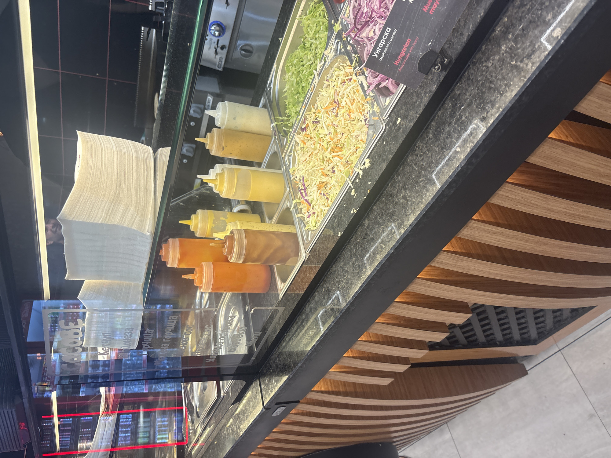 The image shows a food service counter display, likely in Burgas, Bulgaria, featuring various condiments and salad ingredients. In the foreground, a dark granite countertop has several condiment bottles, including orange, yellow, and white sauces, placed in metal trays. To the right, there are trays filled with shredded cabbage and purple onions. A sign in the foreground reads "Унгарска" and "Hungarian," suggesting a specific dish or dressing. Behind the display, a glass partition reveals stacks of white paper napkins. The background shows a dimly lit area with what appears to be beverage refrigerators and kitchen equipment. A person's face is partially visible behind the napkins. The counter itself has a distinctive design with vertical wooden slats.
