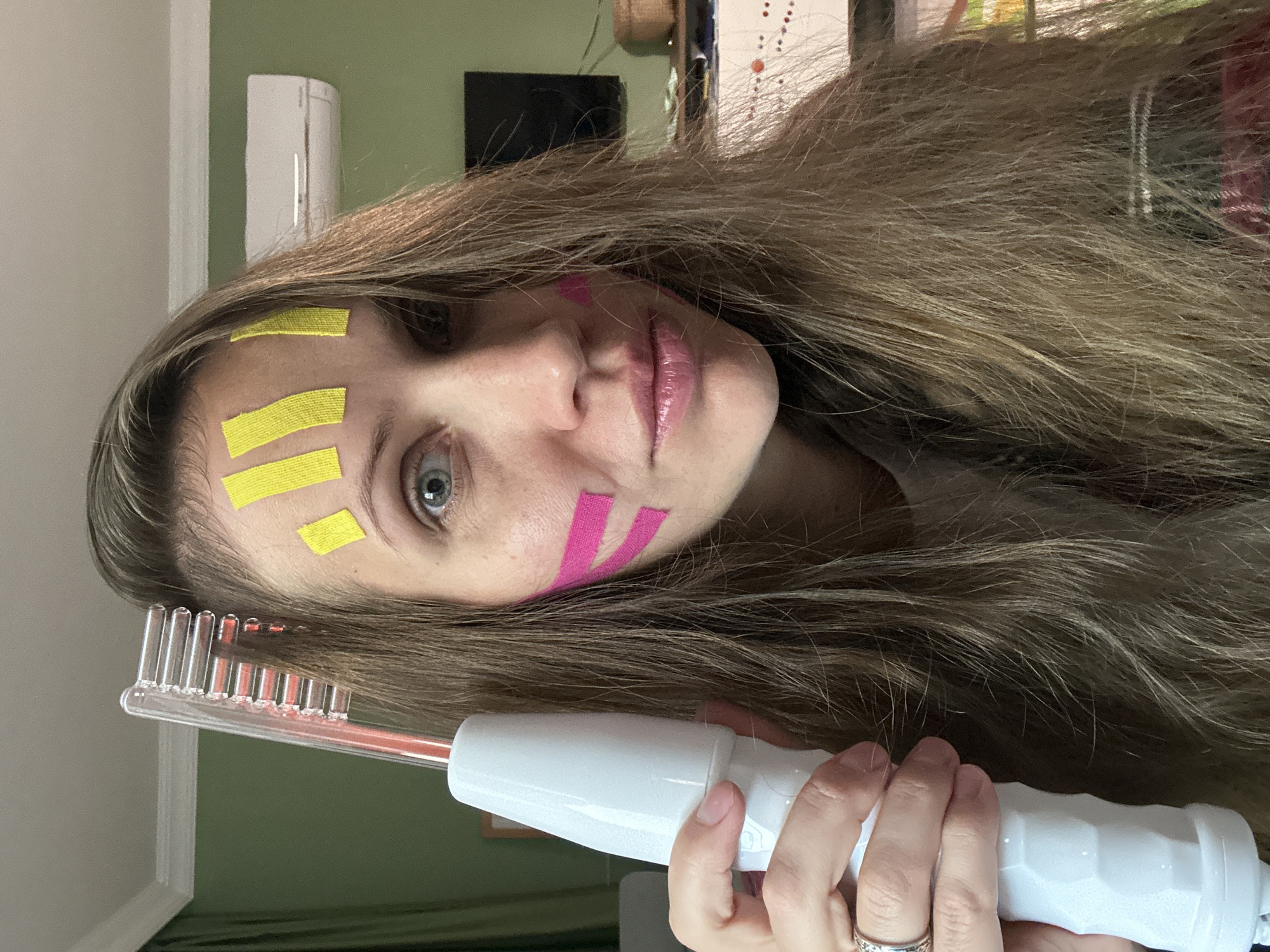 A woman with long brown hair and makeup on her face holds a red comb-like device. There are three yellow stripes on her forehead and two pink stripes on her cheek. She is wearing a silver ring on her finger. The background is a green wall and various objects. The location is not identifiable as Fethiye, Turkey.