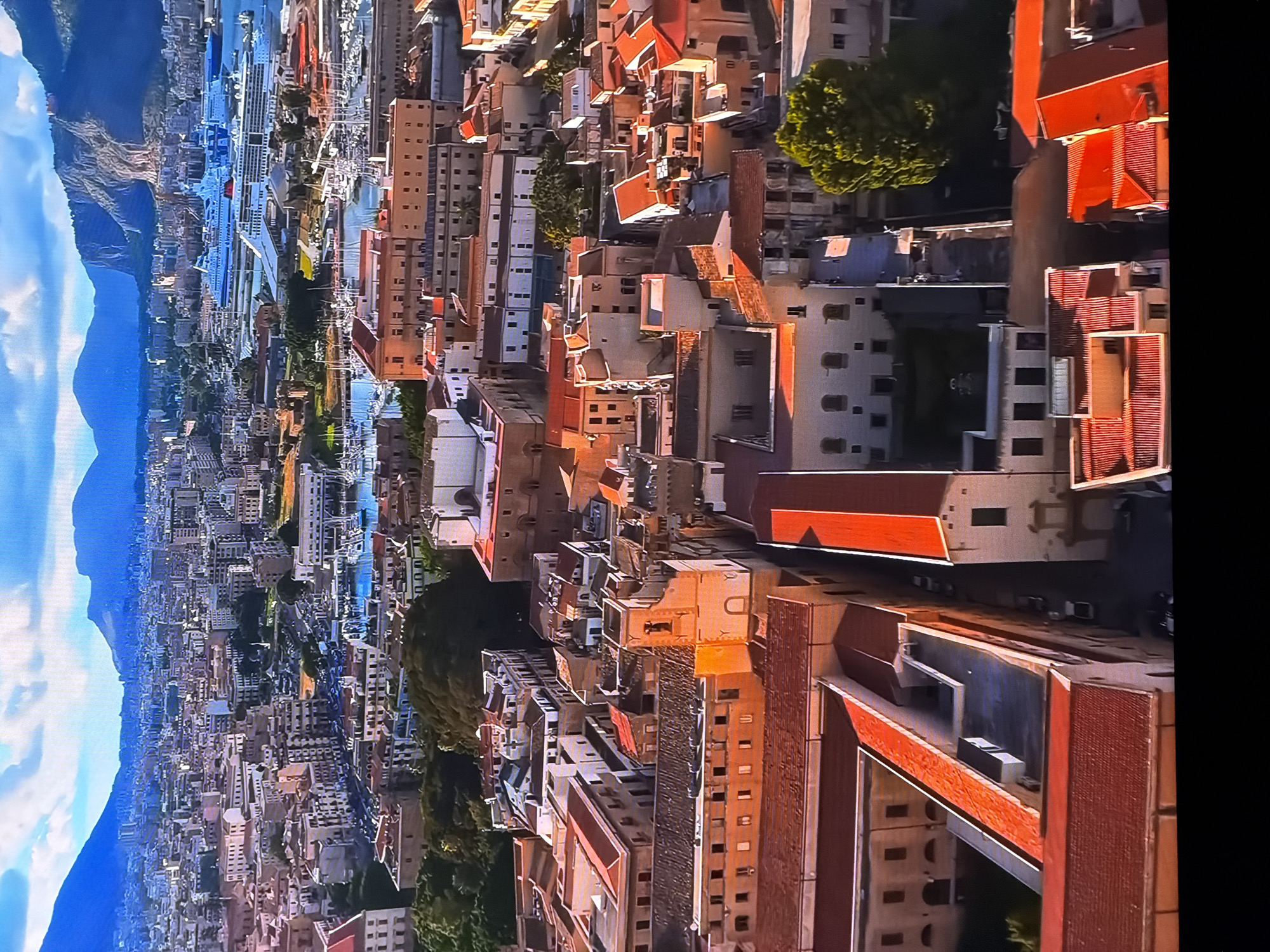 The image displays an aerial view of a densely populated city with a harbor and mountains in the background. The architecture suggests a Mediterranean or European city, characterized by terracotta-tiled roofs, light-colored buildings, and narrow streets. In the foreground, several buildings with red tiled roofs are visible. The middle ground features a bustling harbor with numerous boats and two large cruise ships docked. Beyond the harbor, a vast cityscape extends towards the mountains that form the horizon. The sky is partly cloudy, with patches of blue sky and white clouds. There is no indication of Chirchik, Uzbekistan in this image.