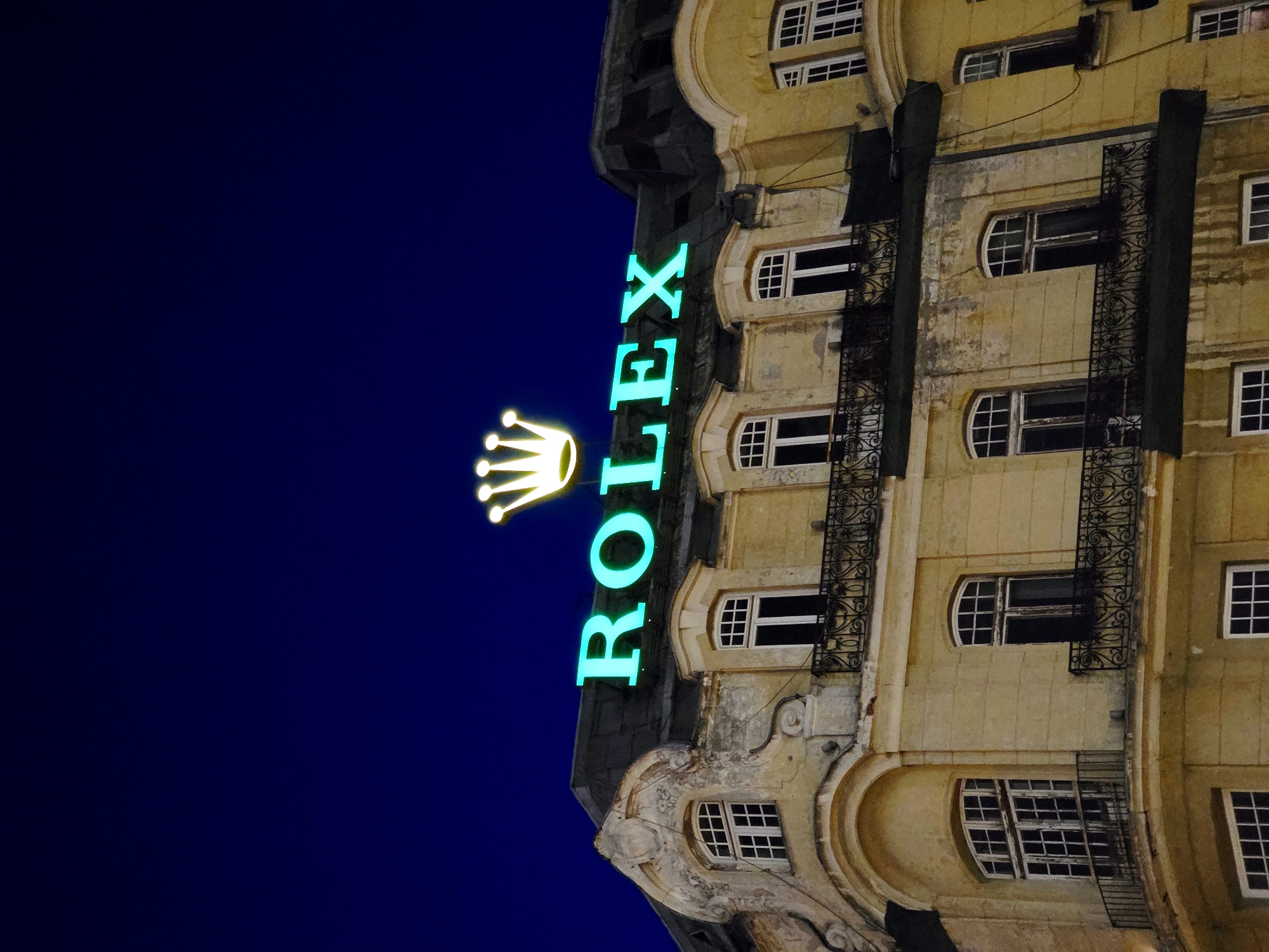 The image shows a nighttime view of a building with a large illuminated "ROLEX" sign and a crown logo on its roof. The building appears to be made of light-colored stone and has multiple windows with balconies. The sky is a deep blue, indicating it is night. The "ROLEX" sign is a vibrant aqua color and is prominently displayed above the roofline. The crown logo, positioned to the left of the text, is illuminated in yellow. The architecture of the building suggests it is an older structure. The image does not provide specific information about the city of Warsaw, Poland, nor does it depict any people, actions, or interactions. The scene is a static, architectural shot focusing on the brand's signage.