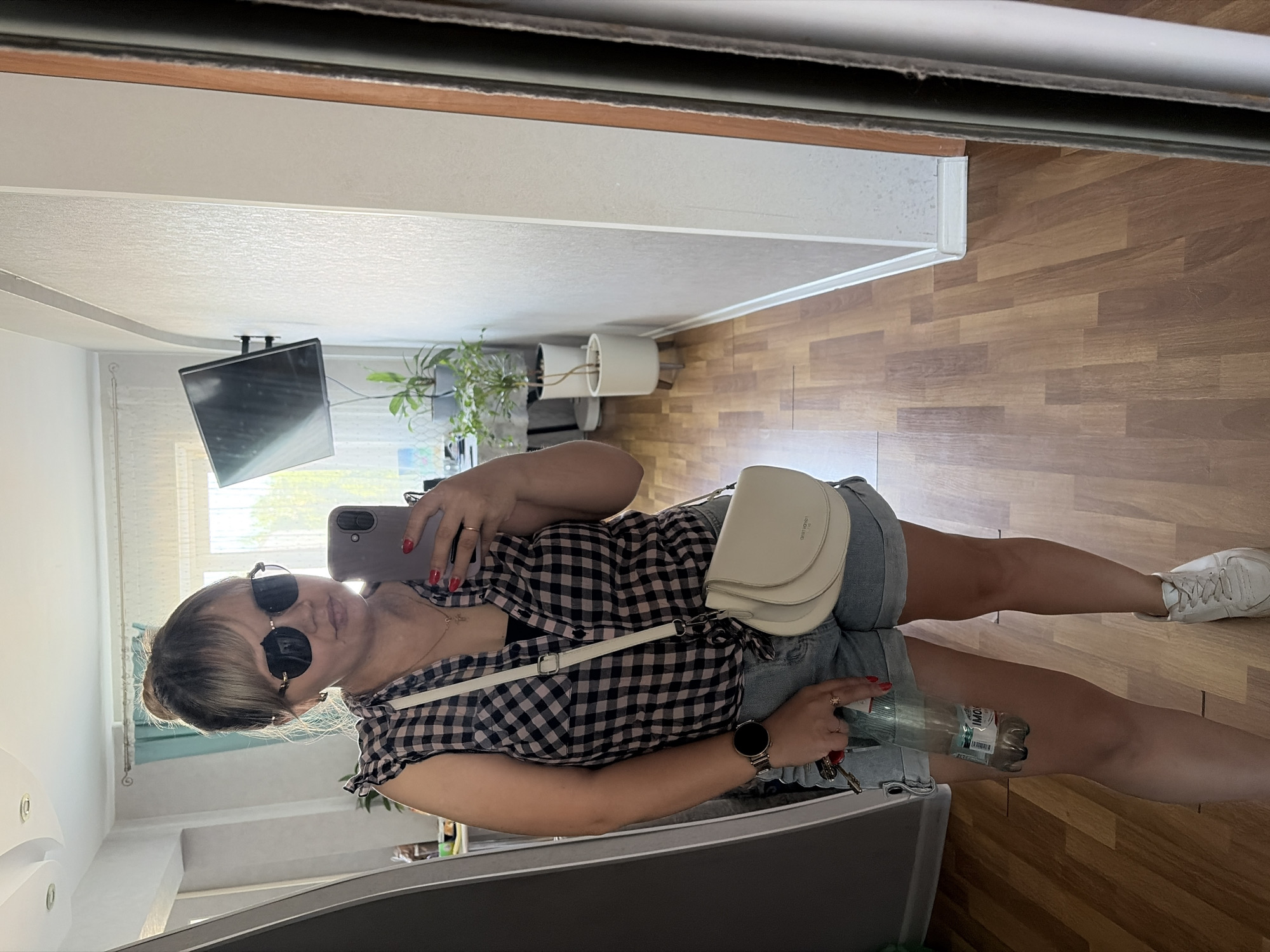 A woman stands inside, taking a selfie in a mirror. She is wearing sunglasses, a plaid shirt, and denim shorts. She has a white purse slung across her body. She is holding a phone in her right hand and a water bottle in her left. She is wearing a watch on her left wrist. Her nails are painted red. The background shows a room with wooden floors, plants in white pots, and a TV mounted on the ceiling. The location may be in Chaikovsky, Russia.