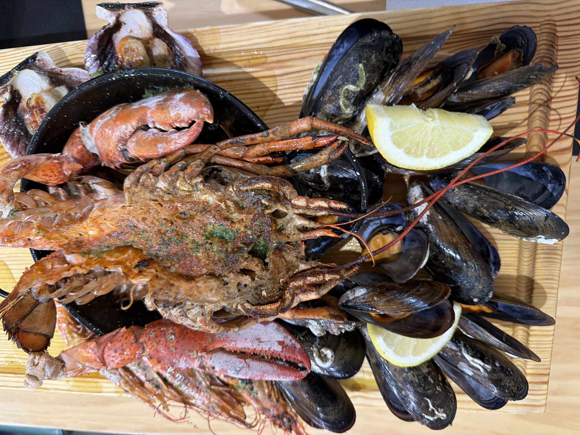 A wooden tray holds a seafood platter, including oysters, prawns, spider crab, lobster and mussels. Two lemon wedges are placed atop the mussels. The platter is likely served in a restaurant in Santiago De Compostela, Spain, based on typical local cuisine.
