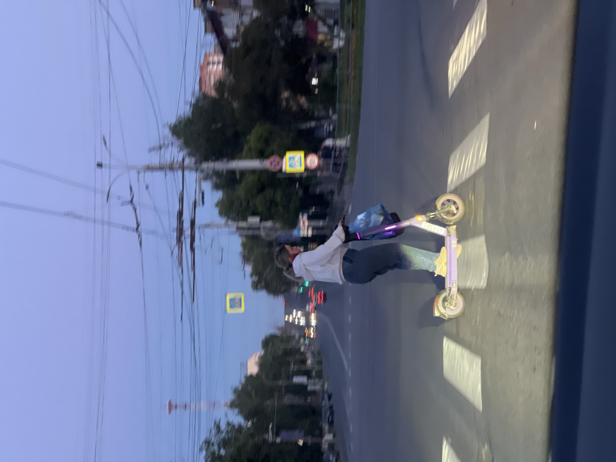 A young woman rides a purple electric scooter across a pedestrian crossing on a street in Izhevsk, Russia. The sky is twilight blue. She is wearing a white hoodie, dark pants, and yellow sneakers, and she is carrying a blue bag. The street is lined with trees and buildings, and there are streetlights and traffic signals visible. Cars are driving in the distance. The pedestrian crossing is marked with white stripes. The scooter has lights on its wheels and frame.