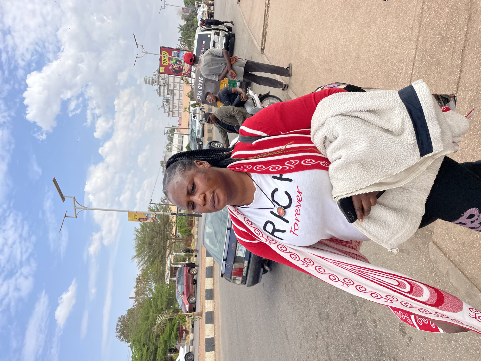 The image shows a woman standing on a sidewalk next to a road in Barakin Lerawi, Nigeria. She is wearing a white shirt with "RICH forever" printed on it, a red and white patterned shawl, a fleece jacket, and black pants. In the background, there are buildings, cars, a tricycle taxi with "PTA-8776" written on it, and a billboard advertising "ye!!". Other people are visible, including a man wearing a red cap and grey shirt, and individuals on the tricycle taxi. The sky is blue with scattered clouds.
