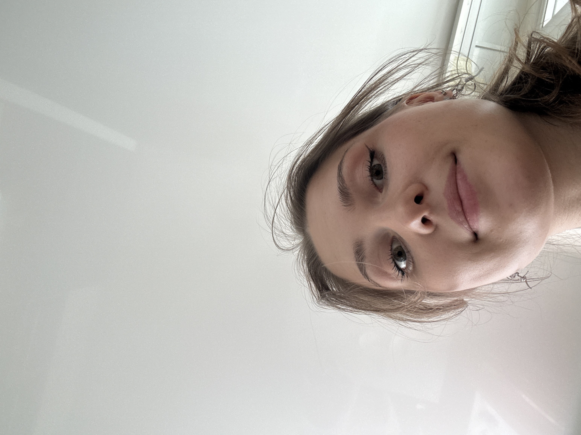 A portrait-oriented photograph captures a close-up of a young woman's face from a low angle, looking upwards. Her head is tilted, and she gazes towards the viewer with a subtle, gentle smile. Her hair is light brown, appearing slightly windswept around her face. Her light-colored eyes reflect the ambient light. A small, decorative silver earring is visible on her left ear. The background consists of a very light, almost white, plain surface, likely an indoor wall or ceiling, featuring some faint reflections or light streaks. The overall setting is brightly lit, suggesting an indoor environment. No other subjects, objects, or specific location cues within Saint Petersburg, Russia, are visible in the minimalist background. There is no visible text. The image conveys a serene and intimate moment.
